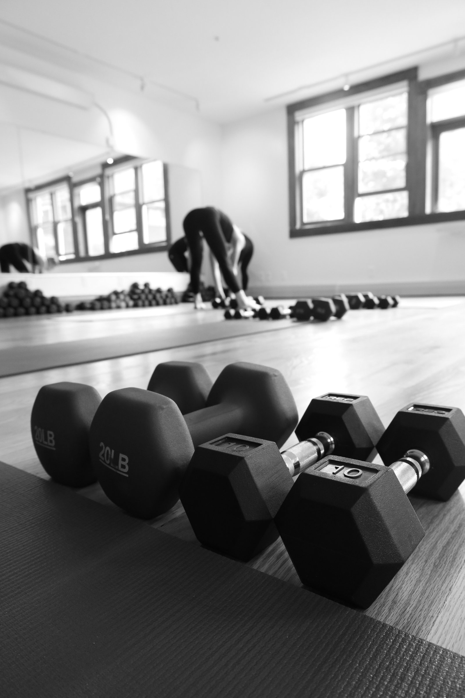Black and white photo of a gym with dumbbells and a person bending down to pick up weights in the background near the windows.