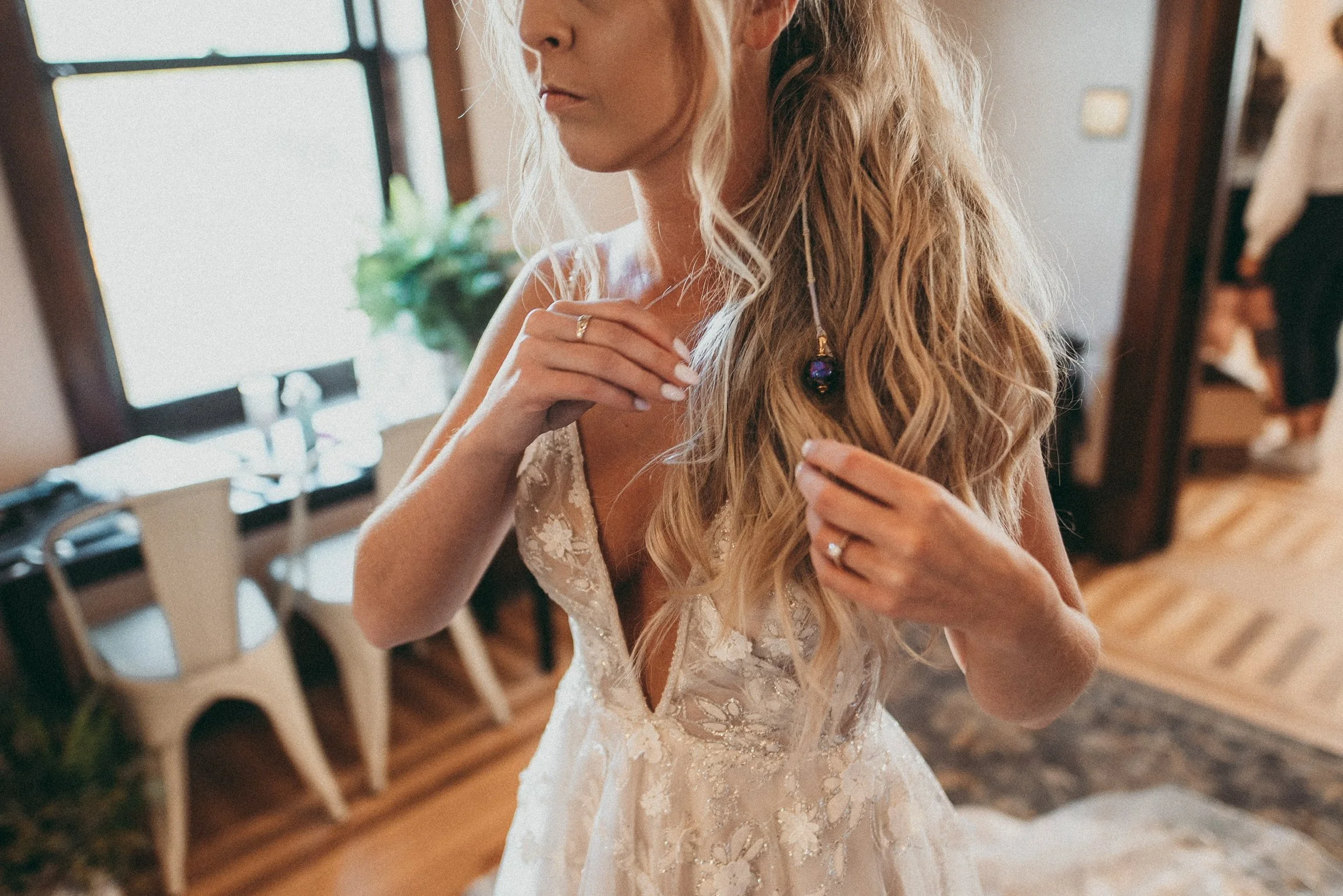 A bride in a wedding dress with lace details is adjusting her hair or jewelry inside a room with wooden floors and furniture. She has long blonde wavy hair and is wearing a ring. She is in a bridal suite