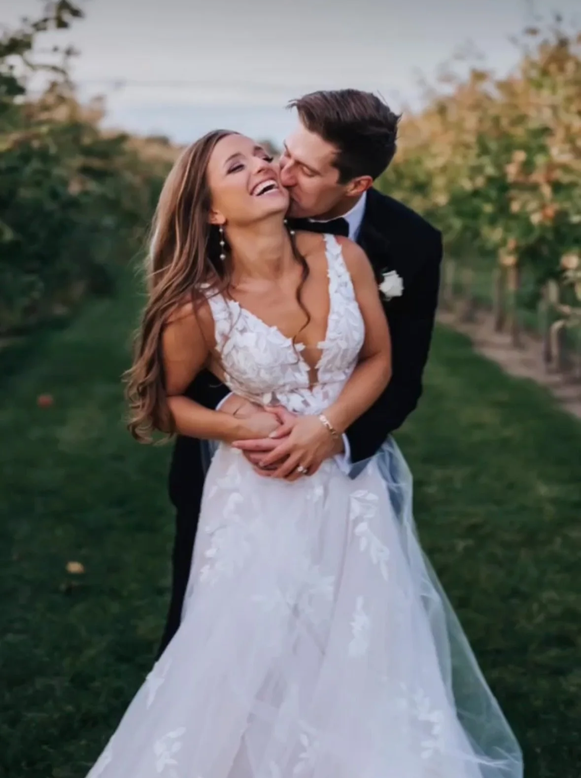 A newlywed couple with a spray tan and soft elegant hair and neutral makeup hugging and sharing a joyful moment in a vineyard, with the groom kissing the bride's cheek. The bride is wearing a white lace wedding dress.