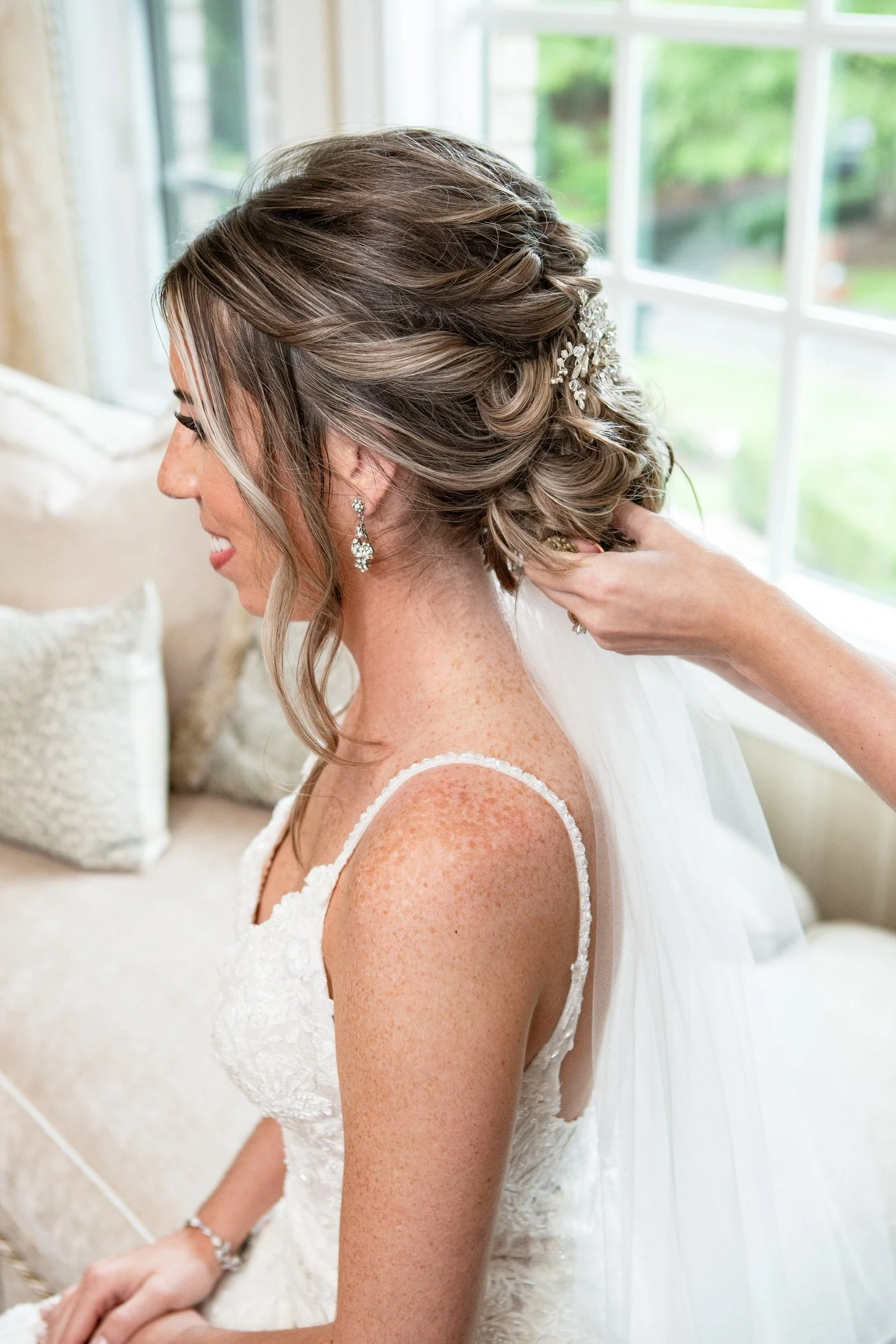 A Hairstylist is doing a brides hair with effortless styled brown hair and a hair accessory, wearing diamond earrings and a sleeveless lace wedding dress, is adjusting her jewelry in a bright room with large windows and green outdoor scenery.
