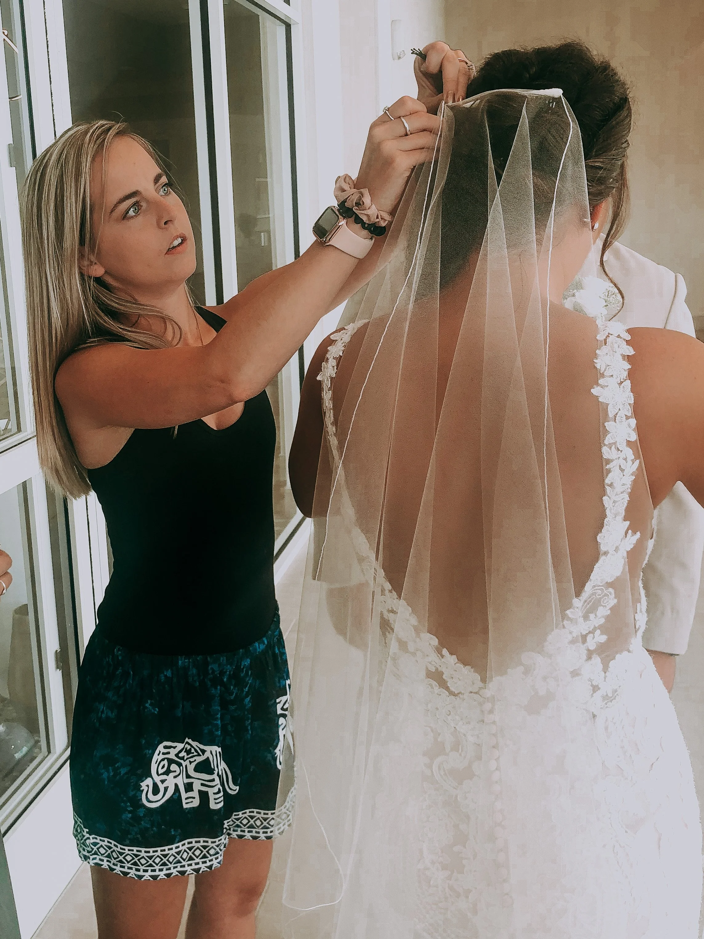 A Hairstylist  is securing a bridal veil on a bride, who is wearing a white lace wedding dress with floral details on the straps.