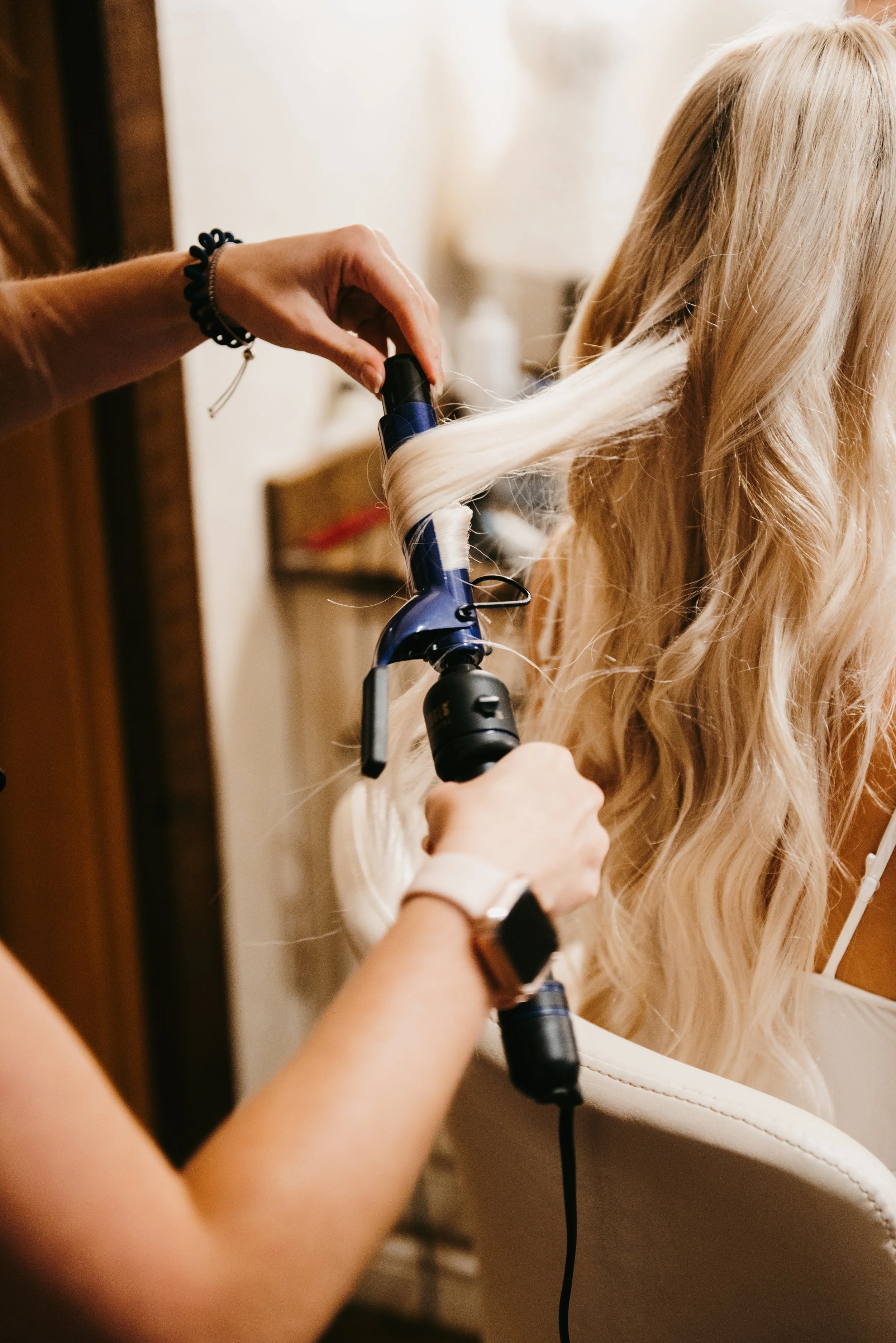 A Hairstylist is curling a woman's long, blonde hair with a curling iron in a salon or home setting.