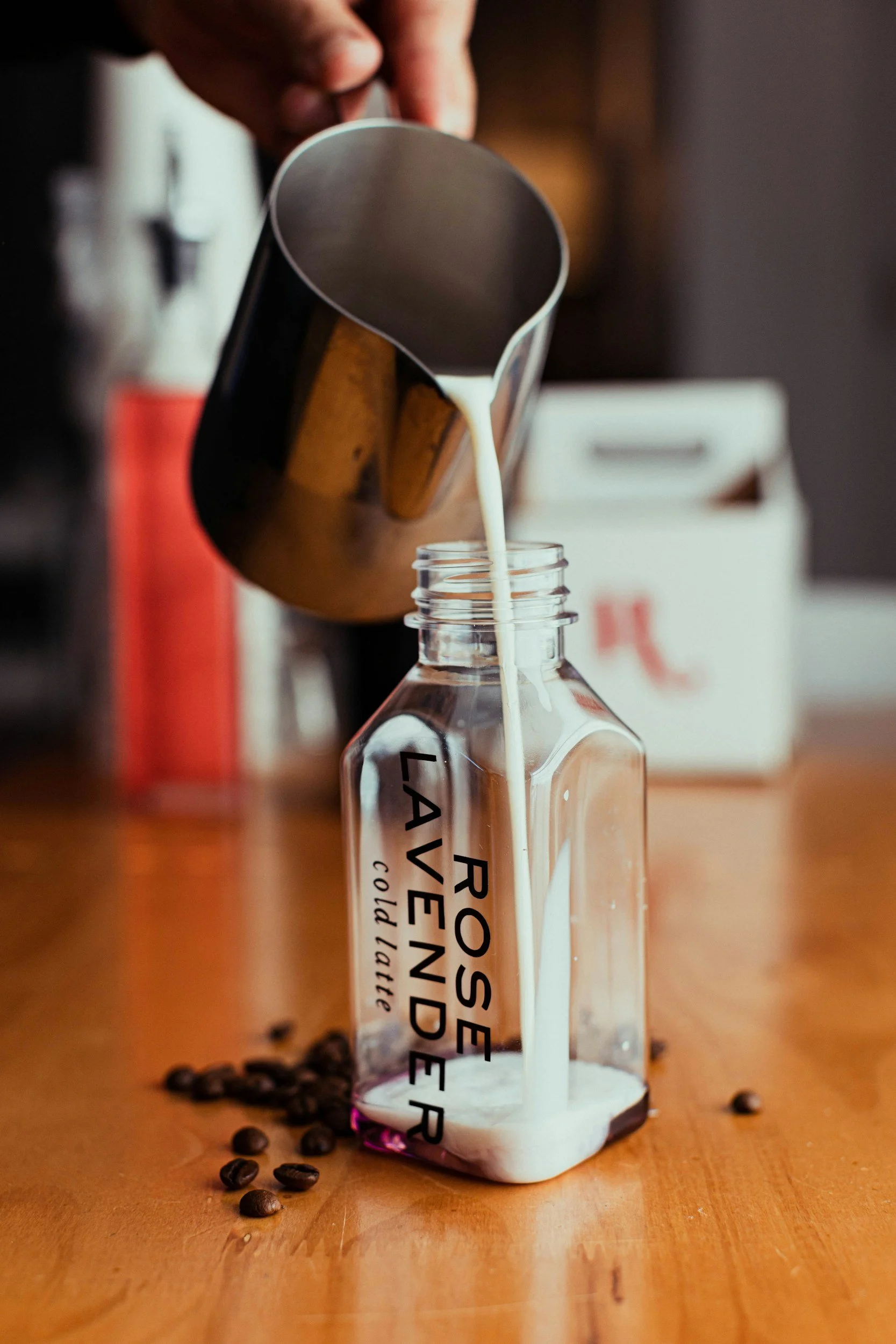 Milk being poured from a metallic pitcher into a clear glass bottle labeled 'Provence Lavender Cold Air.' Coffee beans scattered on a wooden surface around the bottle.