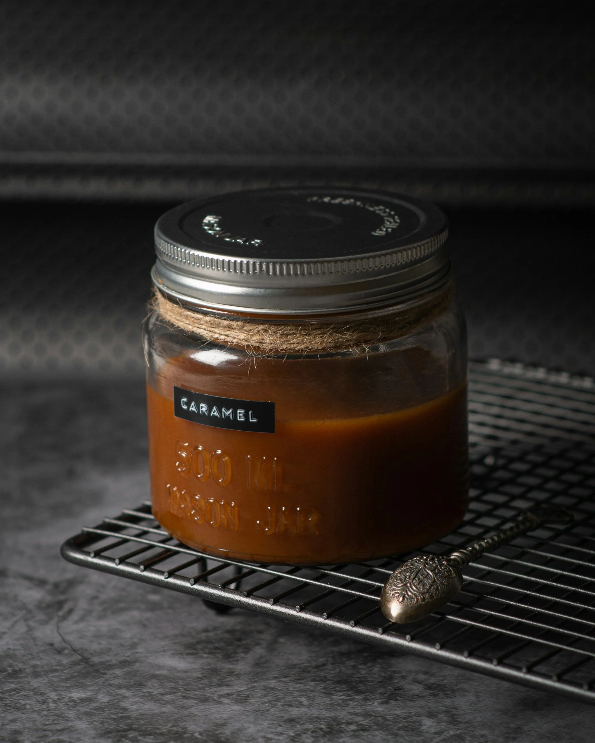 A jar of caramel spread placed on a wire cooling rack with a vintage spoon beside it, against a dark background.
