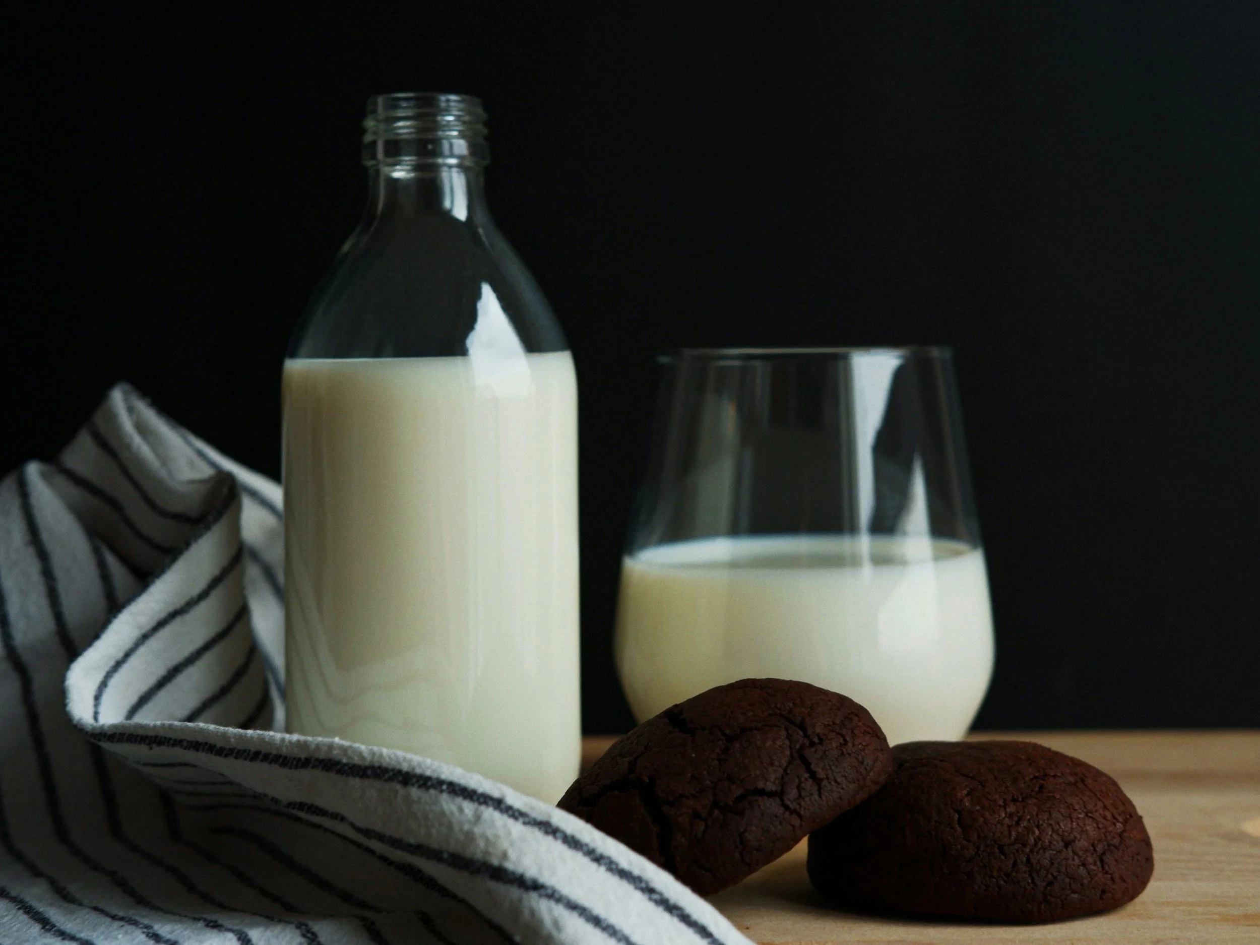 A glass bottle of milk, a glass filled with milk, and two chocolate cookies on a wooden surface with a dark background.