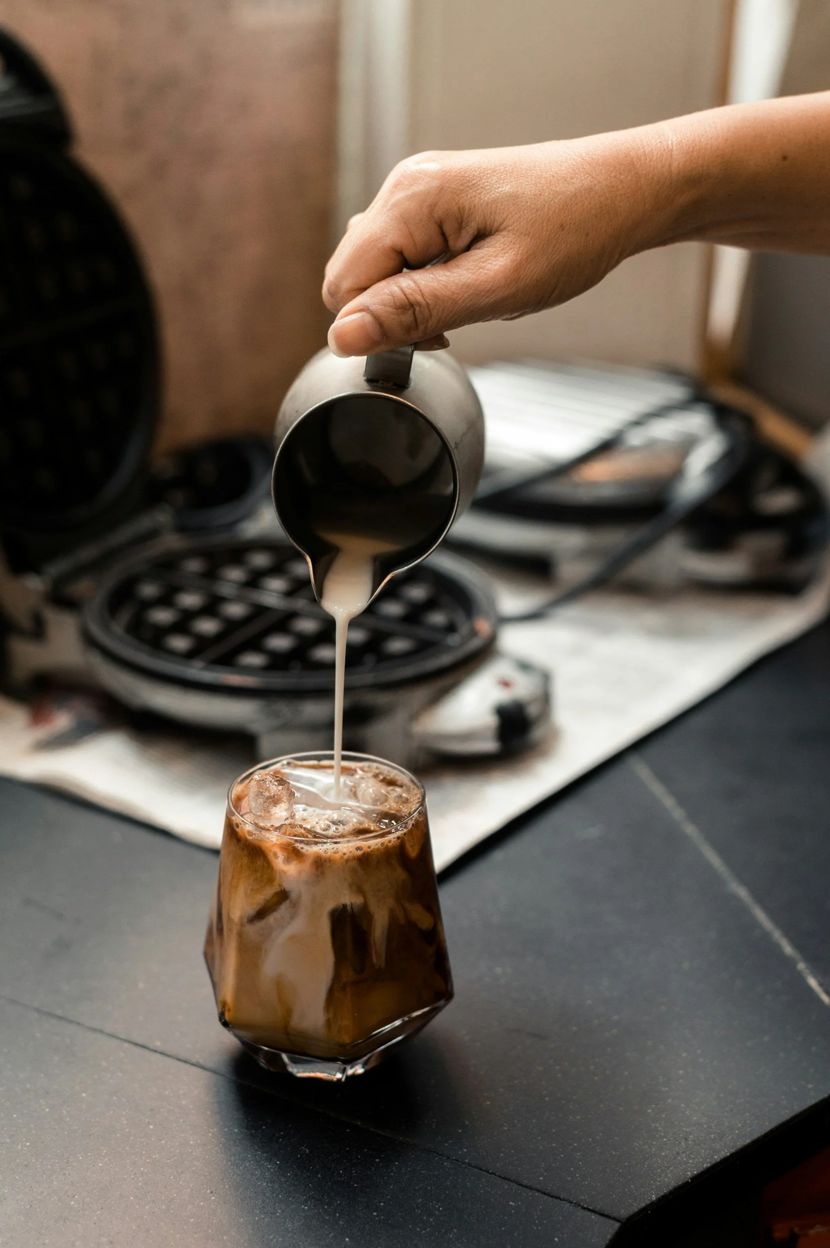 A person's hand pours coffee from a stainless steel pitcher into a glass filled with ice and milk, with a coffee machine in the background.