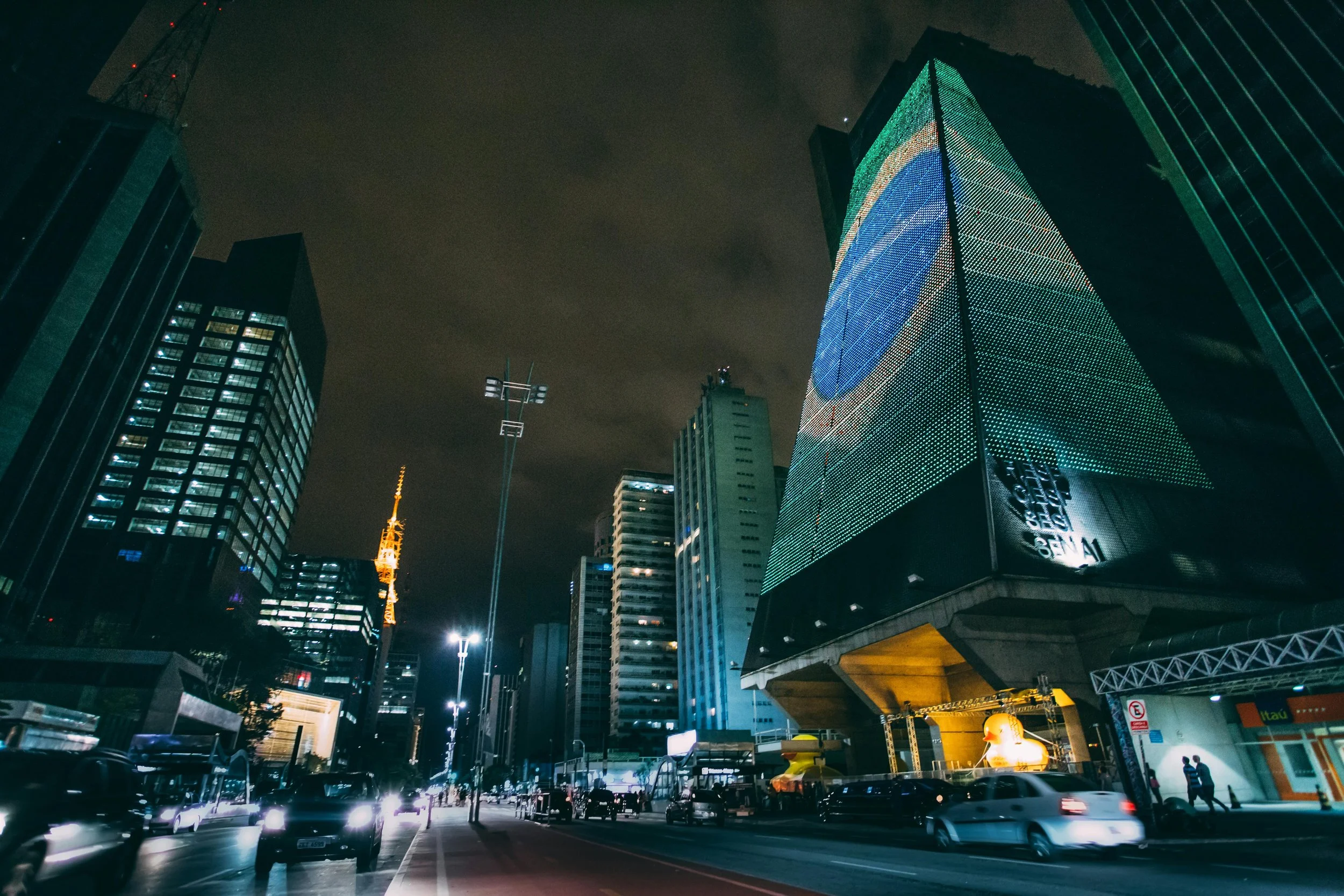 Nighttime cityscape featuring illuminated high-rise buildings with digital billboards, traffic moving on the street, and the sky obscured by clouds.