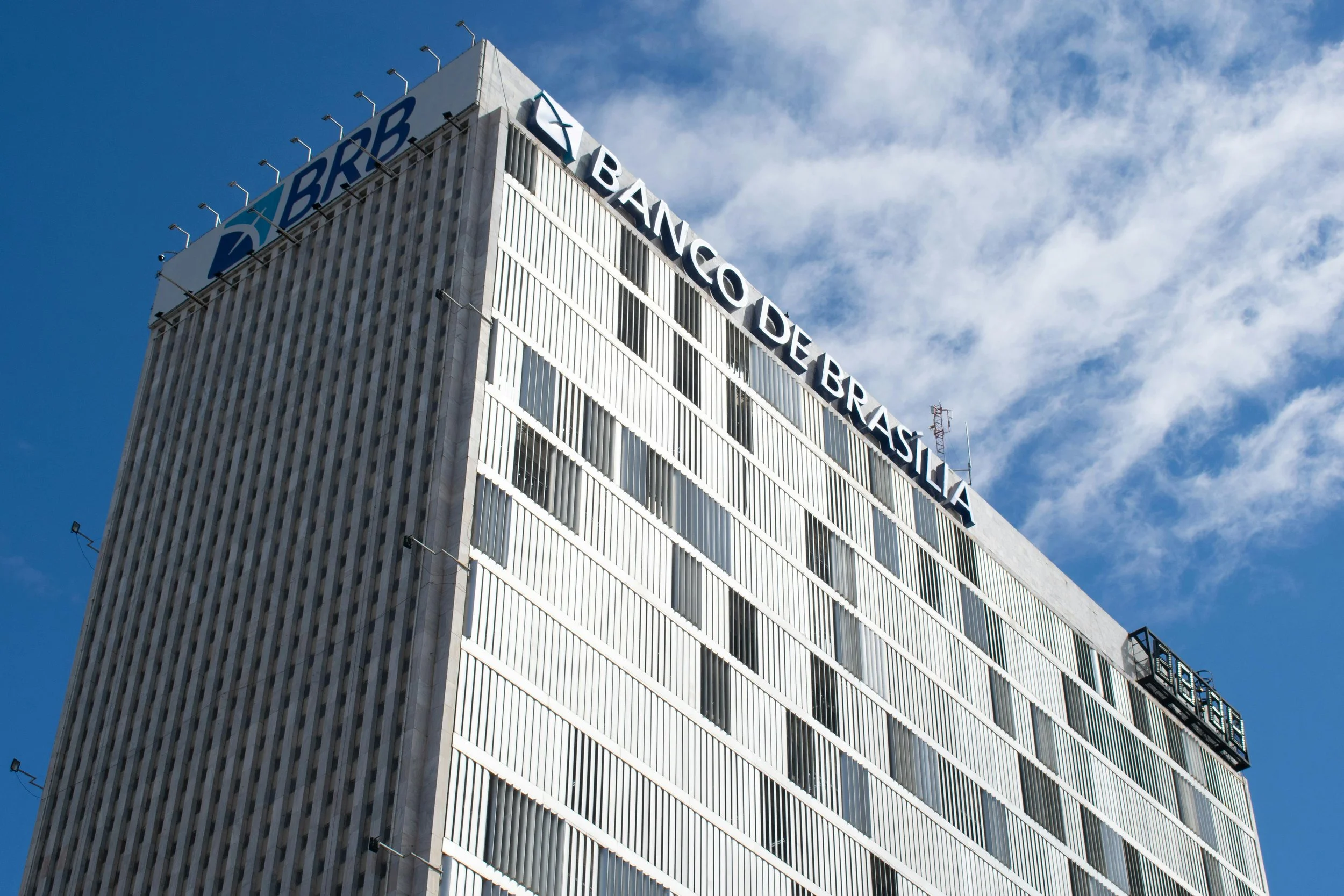 A high-rise building with the logo and name of Banca de Brasília on top. The building has a modern facade with vertical white lines and many windows. The sky is partly cloudy with some blue visible.