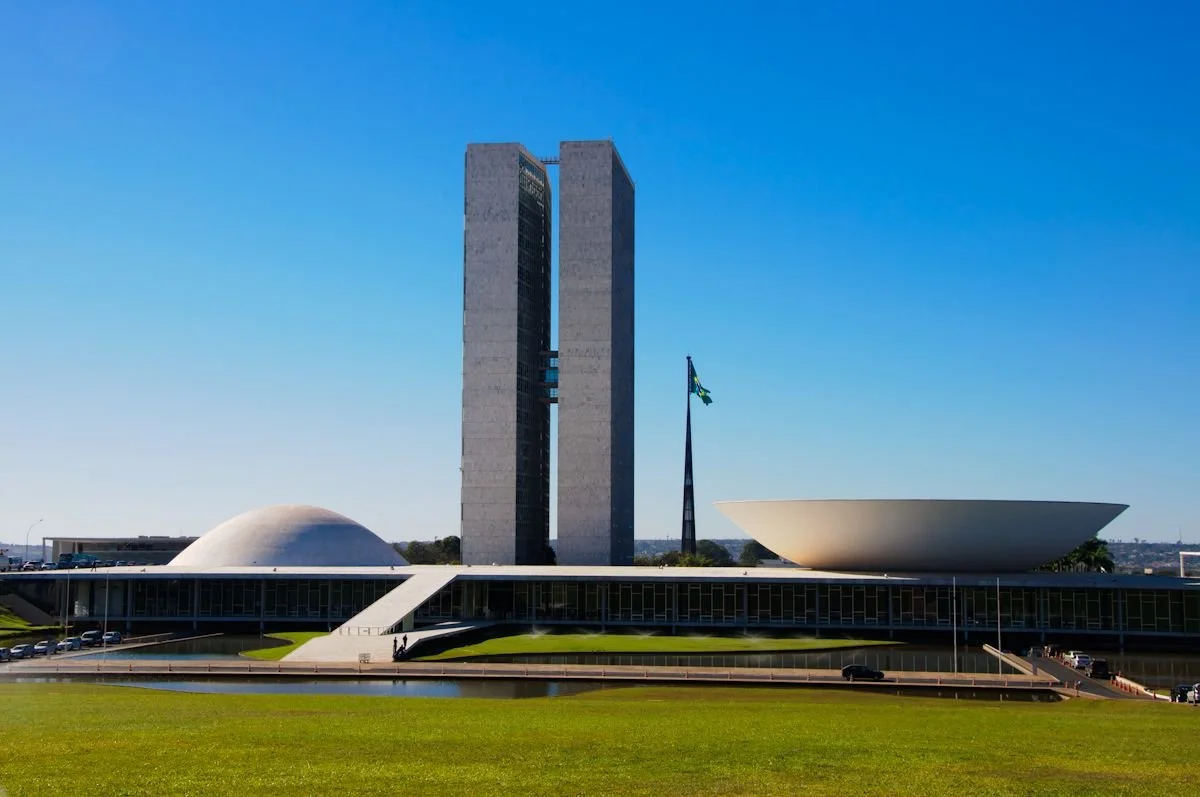The National Congress of Brazil, a modern government building in Brasília with two tall towers, a dome, and a large bowl-shaped structure, against a clear blue sky.