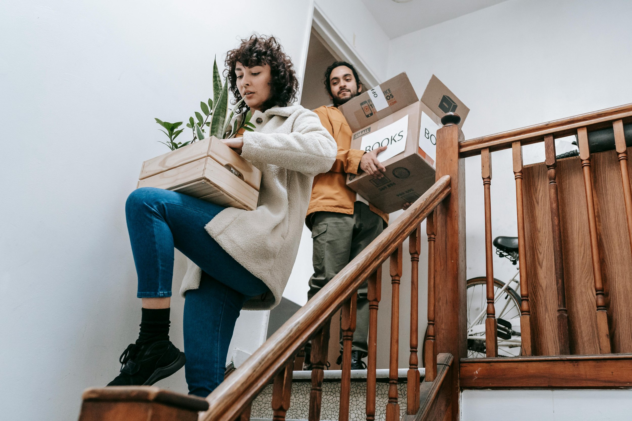 Two people carrying boxes down a staircase, one woman sitting on the stairs arranging a plant, and one man carrying a box labeled 'Books'.