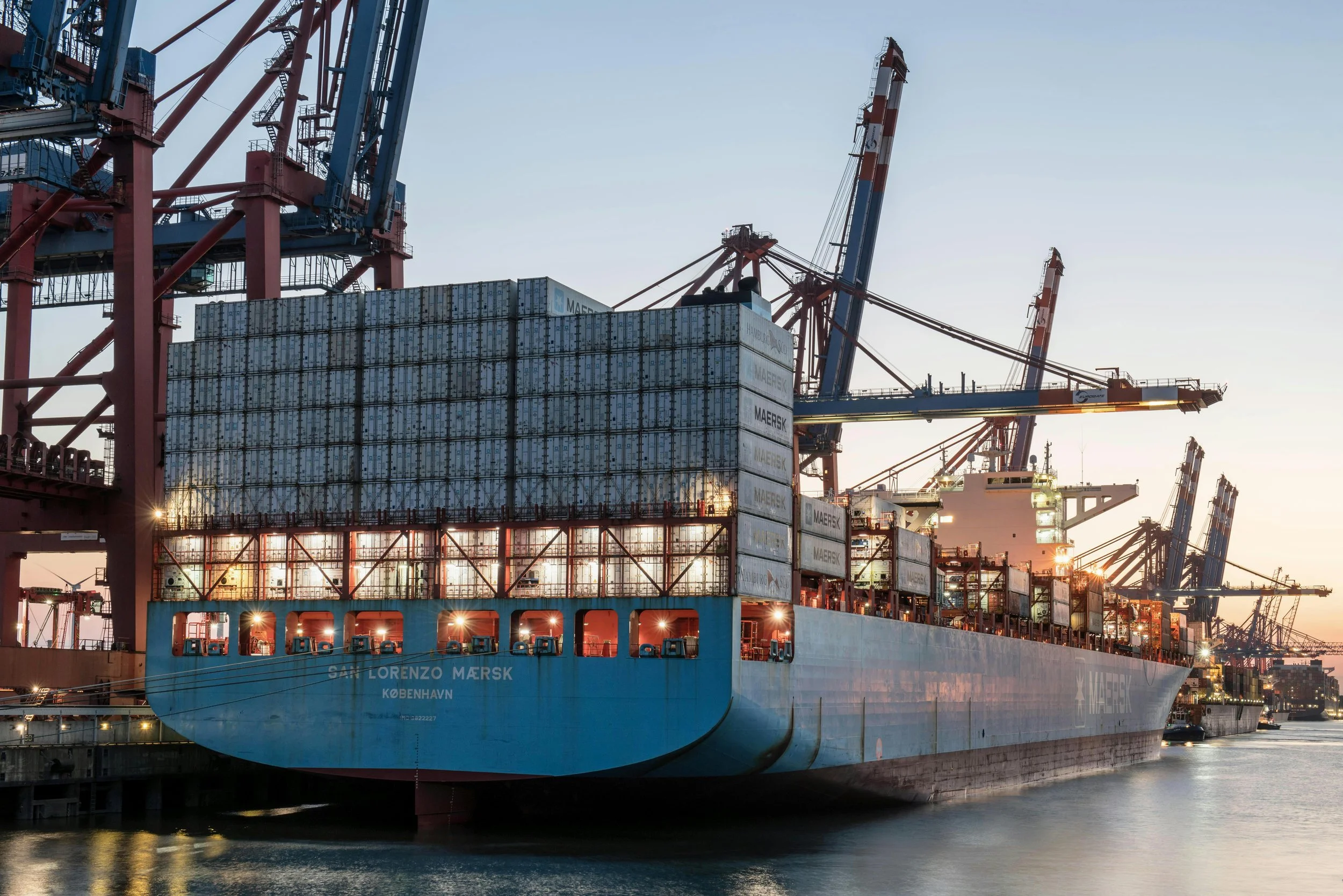 A cargo ship labeled 'San Lorenzo Mersk' docked at a port with large cranes overhead and stacked containers on board, during sunset.