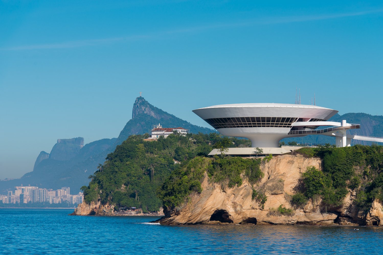 Famous modern building on a hillside by water, with mountains and the Christ the Redeemer statue in the background, in Rio de Janeiro, Brazil.