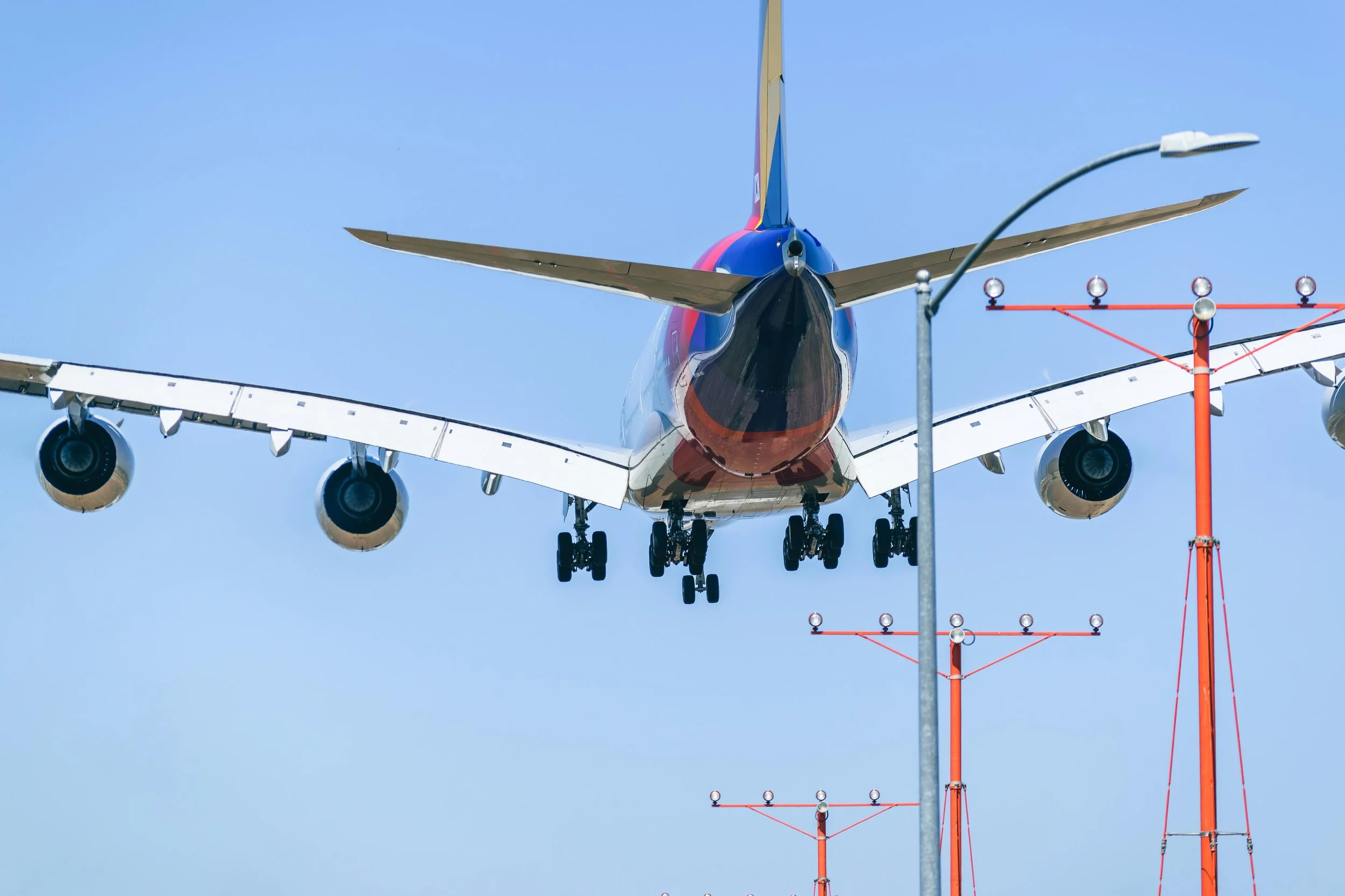 A commercial airplane flying low towards the runway, with an air traffic control tower's lights and antenna in the foreground and a clear blue sky.