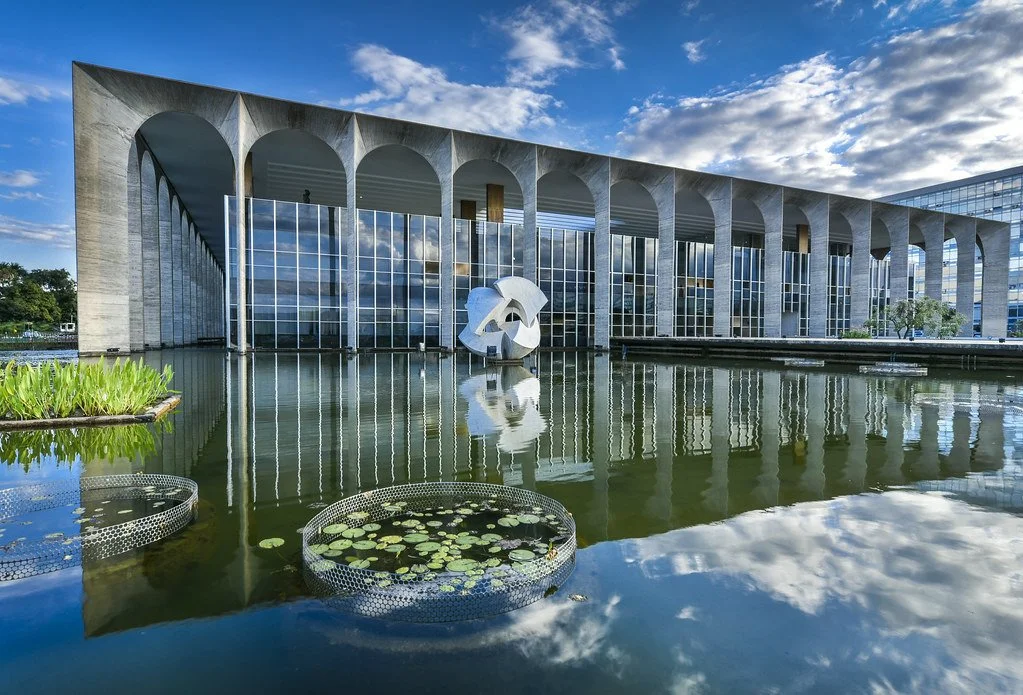 Modern building with large arched windows and concrete arches across the top, reflected in a surrounding pond with lily pads and aquatic plants.