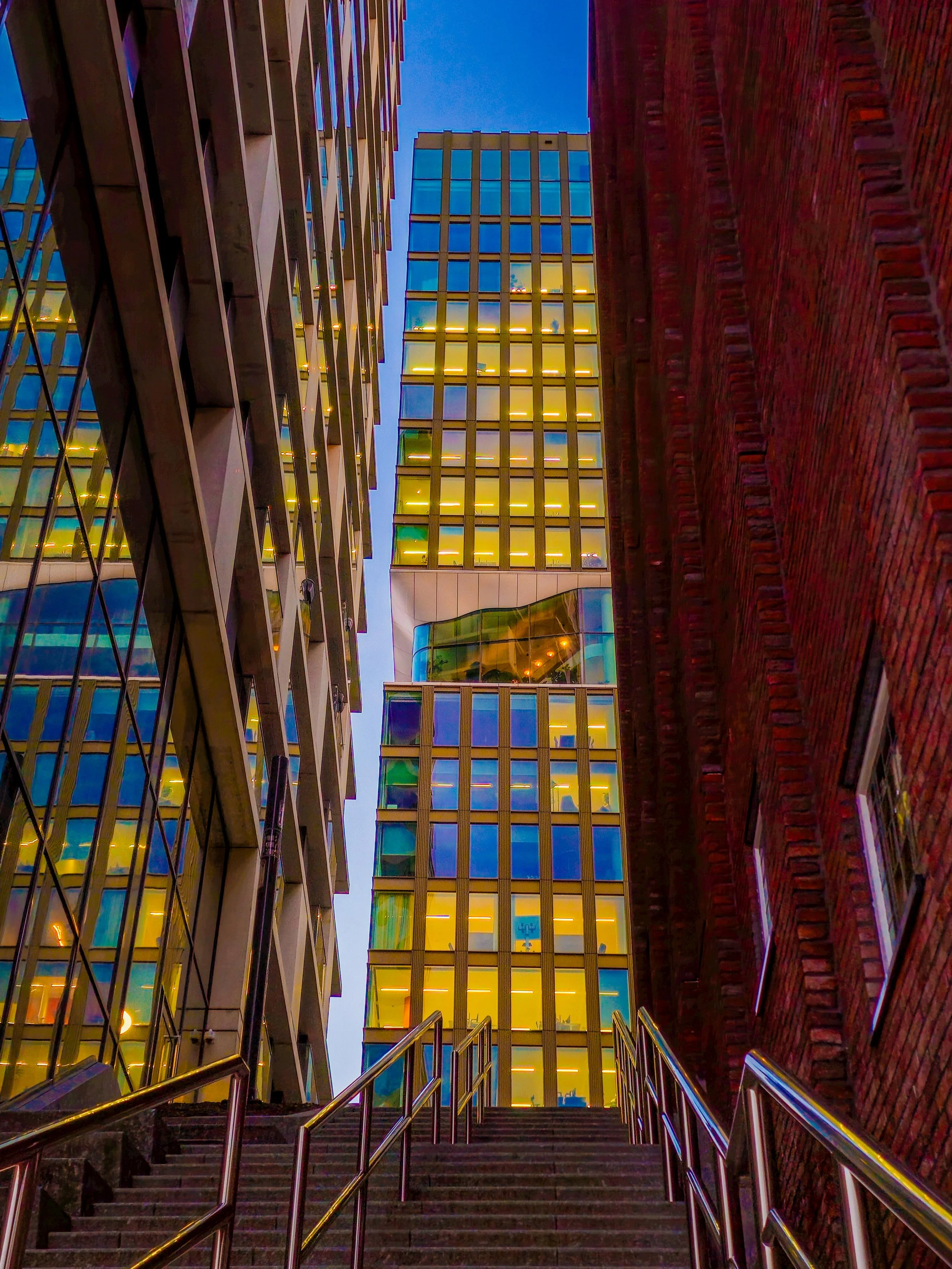 Low-angle view of modern buildings with glass windows and brick walls, with stairs and handrails leading up towards the sky at dusk.