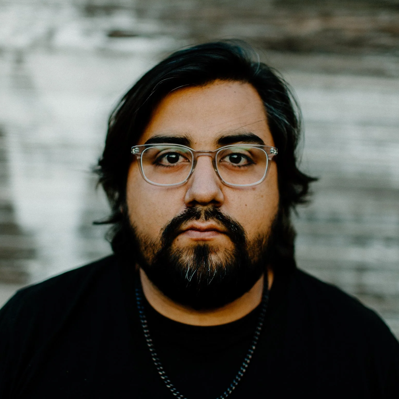 A man with shoulder-length dark hair, glasses, and a beard, looking directly at the camera against a blurred background.