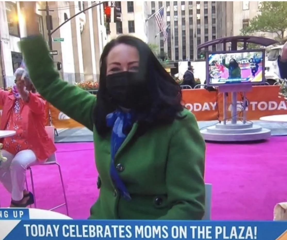 Alli McGill on TV as Today Show Mom of the Year wearing a black face mask and green coat, raising her hand in celebration at an outdoor event on the plaza, with a television camera and pink carpet in the background.