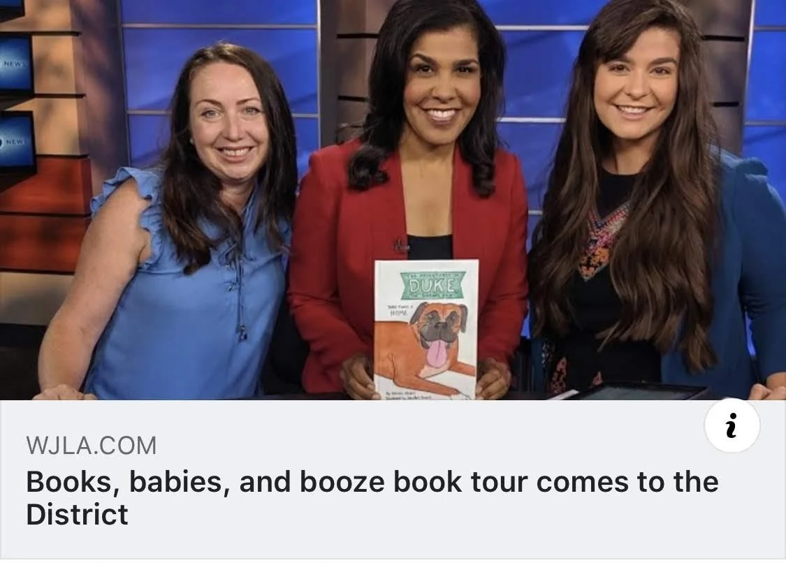 Three women standing together in a studio, holding a children's book titled 'Duke' with a picture of a dog on the cover.