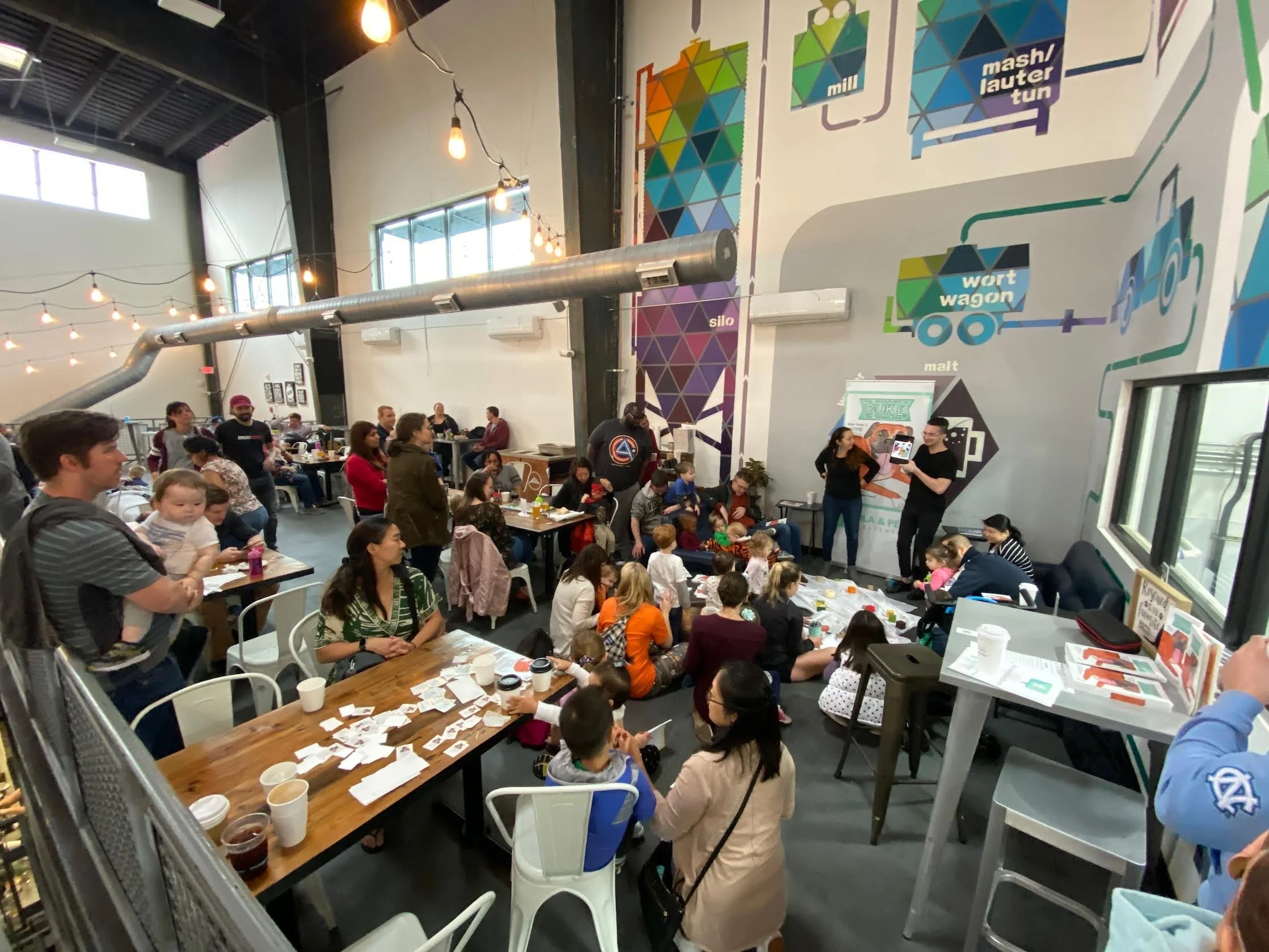 A large indoor book signing and reading event with many people sitting and standing at tables, engaging in an event or workshop, with colorful geometric wall murals and string lights hanging from the ceiling.