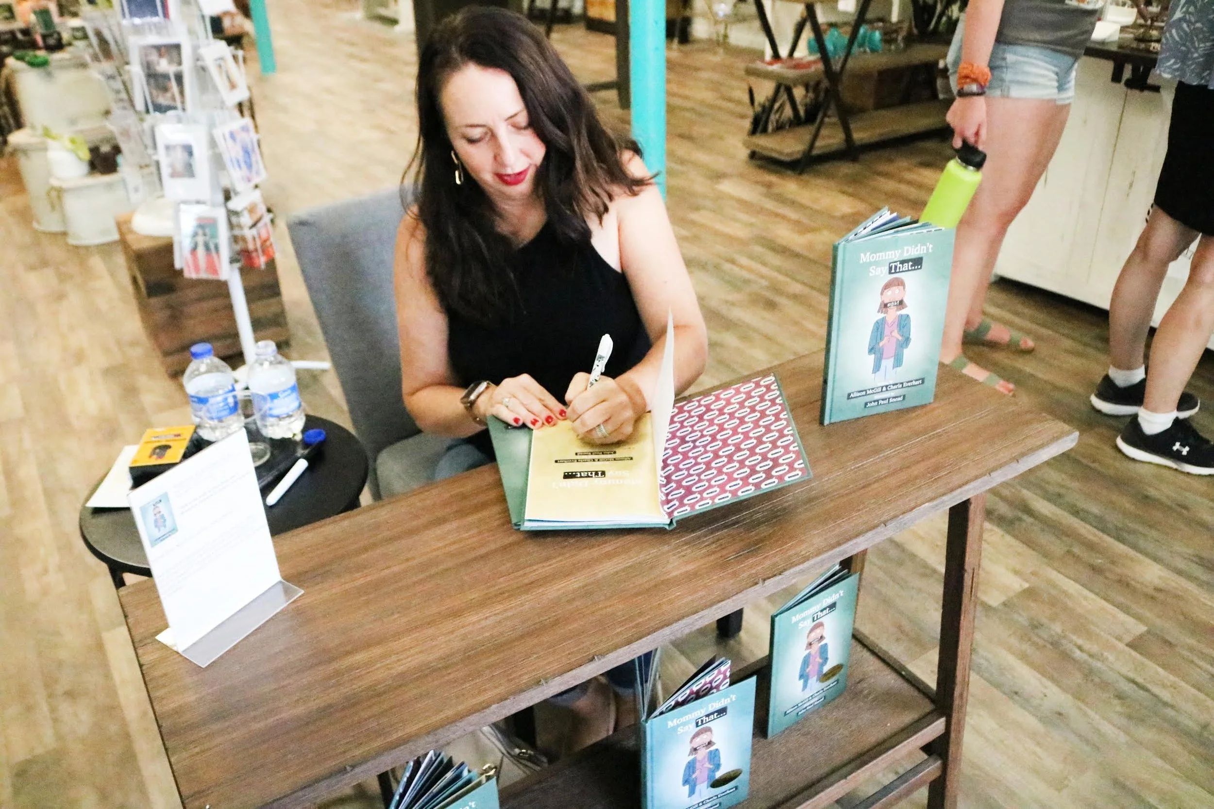 Alli McGill sitting at a table signing a book titled 'Mommy Didn't Say That...' at a book signing event. There are water bottles and promotional material on the table, with additional books displayed nearby.