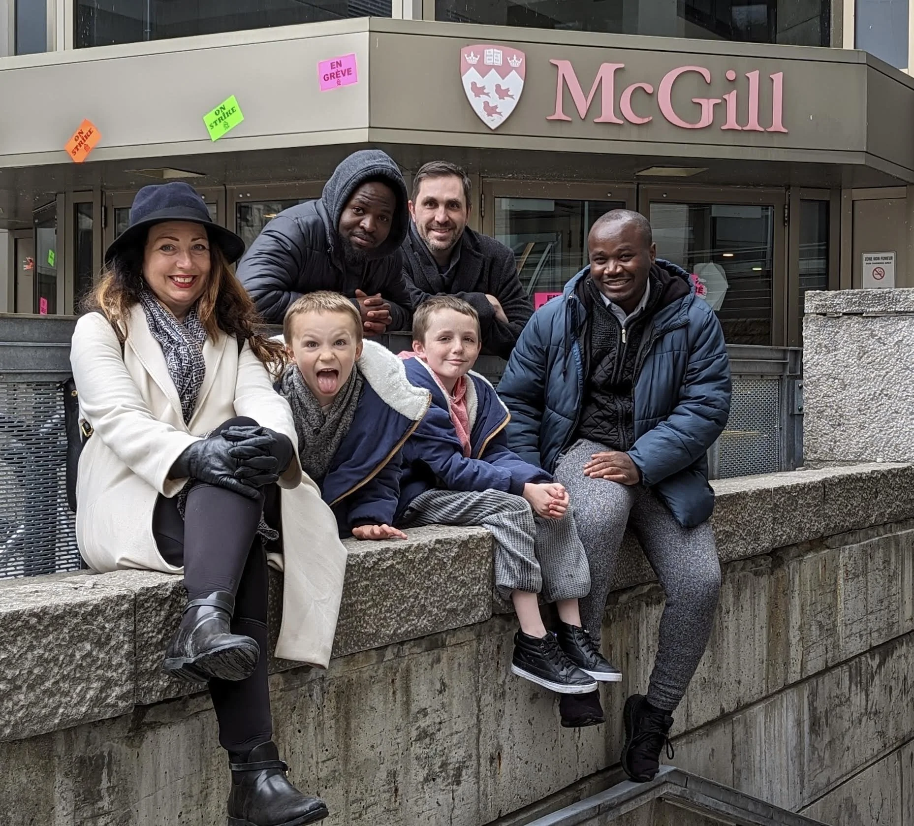 The McGill family, featuring Alli and Joe with their 4 kids, sitting and standing on a stone ledge outside of McGill University building. The background shows the McGill sign and university shield, with colorful protest signs.
