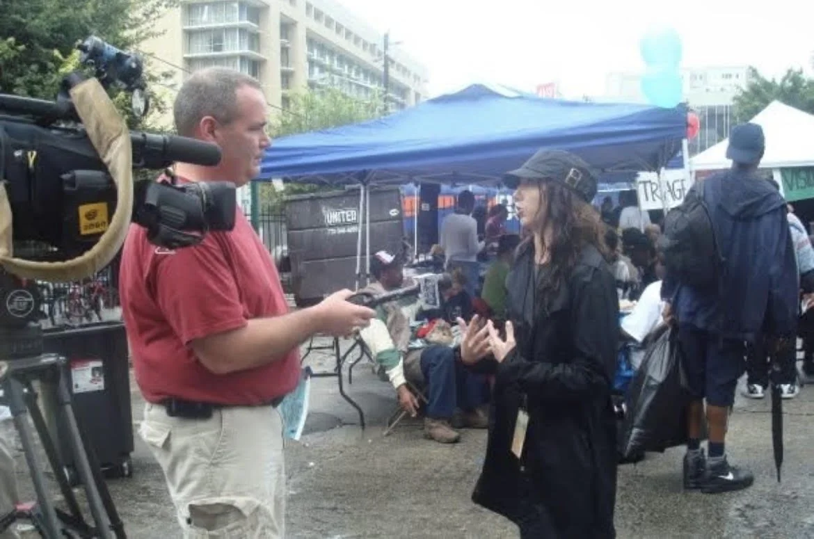 A news reporter interviewing a woman at an outdoor event with tents and people in the background.