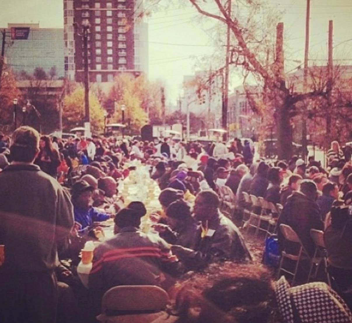 A large outdoor gathering with many people seated at long tables, possibly at a community event or festival, with a cityscape and trees in the background.