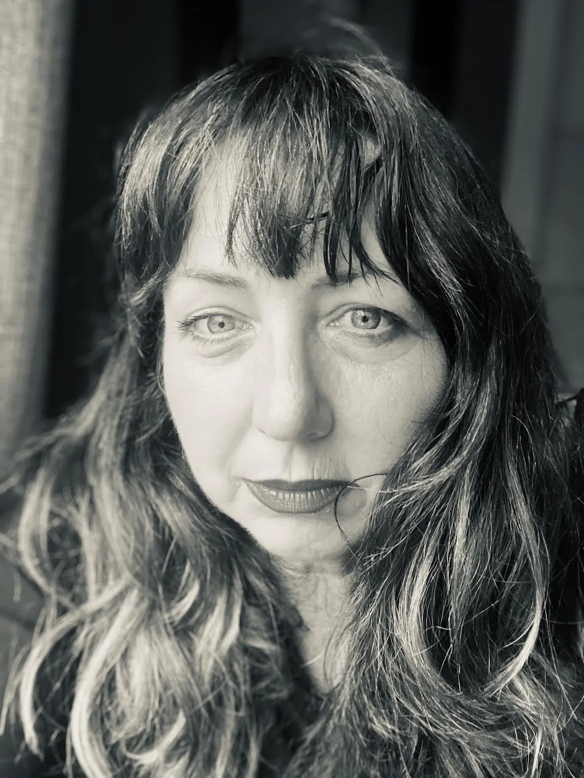 Headshot of Alli McGill. A close-up black and white portrait of a woman with long wavy hair, light eyes, and makeup, looking directly at the camera.