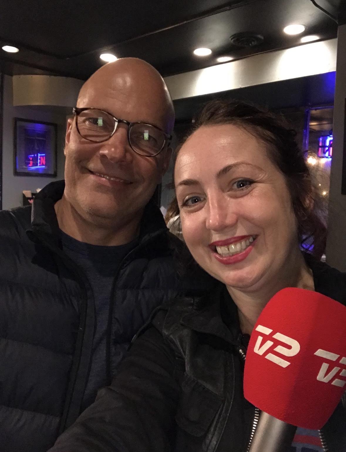 A man and woman taking a selfie together inside a dimly lit room, with a red microphone labeled 'TV 12' in the foreground, both smiling at the camera.
