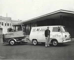 A man standing next to a white van with business branding, and an image of a pickup truck attached to a trailer, parked in front of a building with modern architecture.