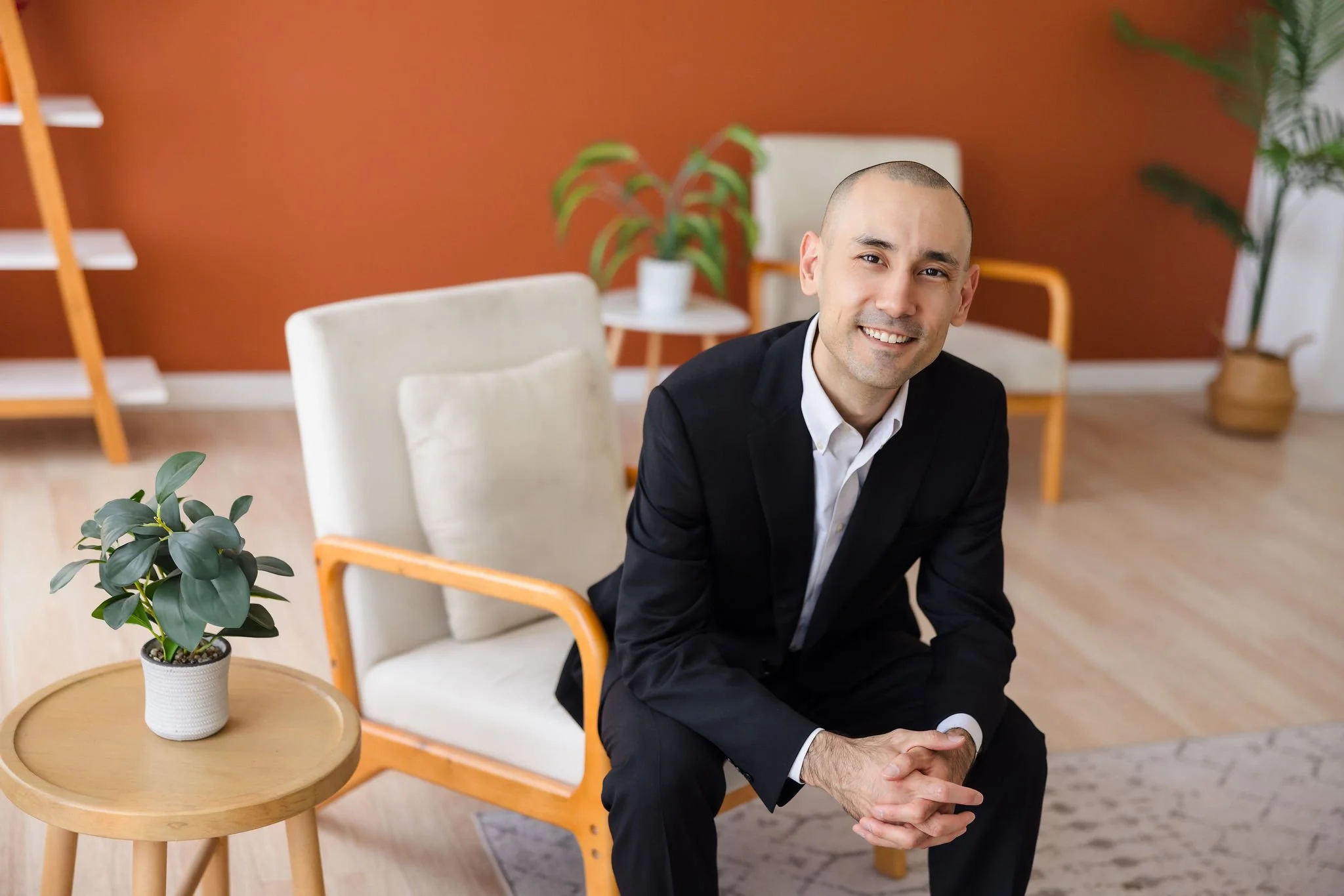 Dr. Riccardi-Zhu is wearing a black suit and white button-up shirt, sitting and smiling at the viewer.