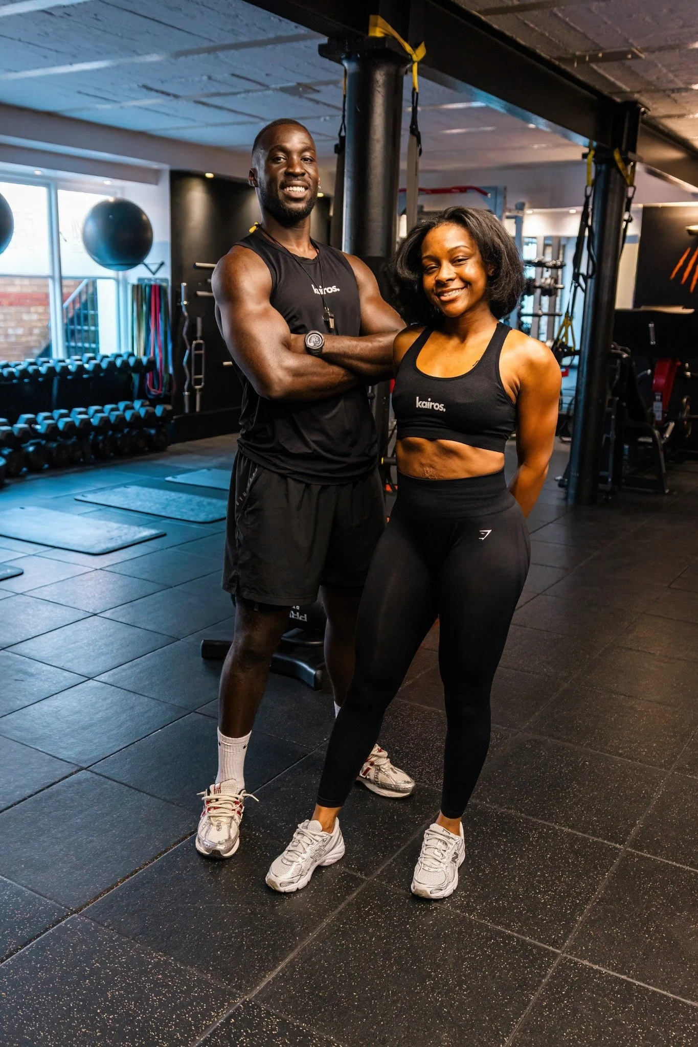 Two fit individuals, a man and a woman, pose in a gym. The man is muscular, wearing a black sleeveless shirt and shorts, with arms crossed. The woman is also athletic, wearing a black sports bra and leggings, smiling with hands behind her back. The gym has black flooring, with exercise equipment visible in the background.