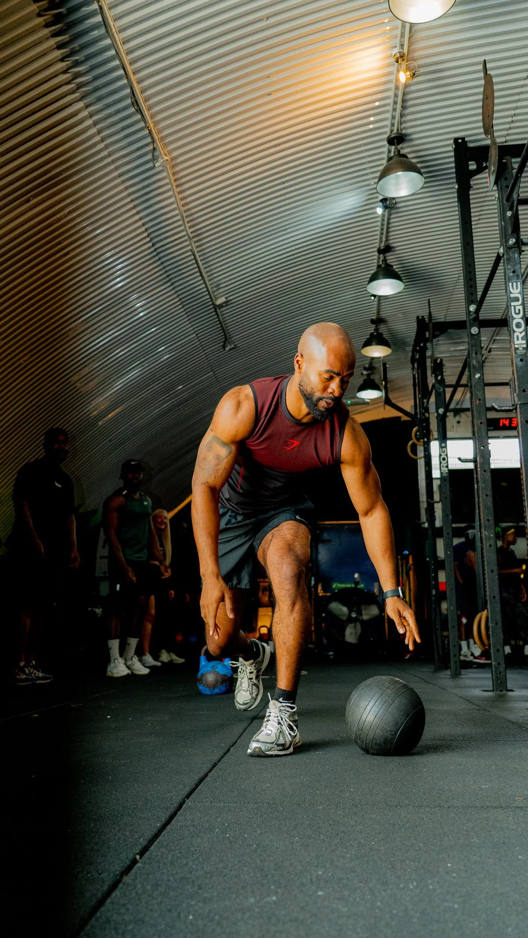 A man performing a medicine ball exercise in a gym, with other people and fitness equipment visible in the background.
