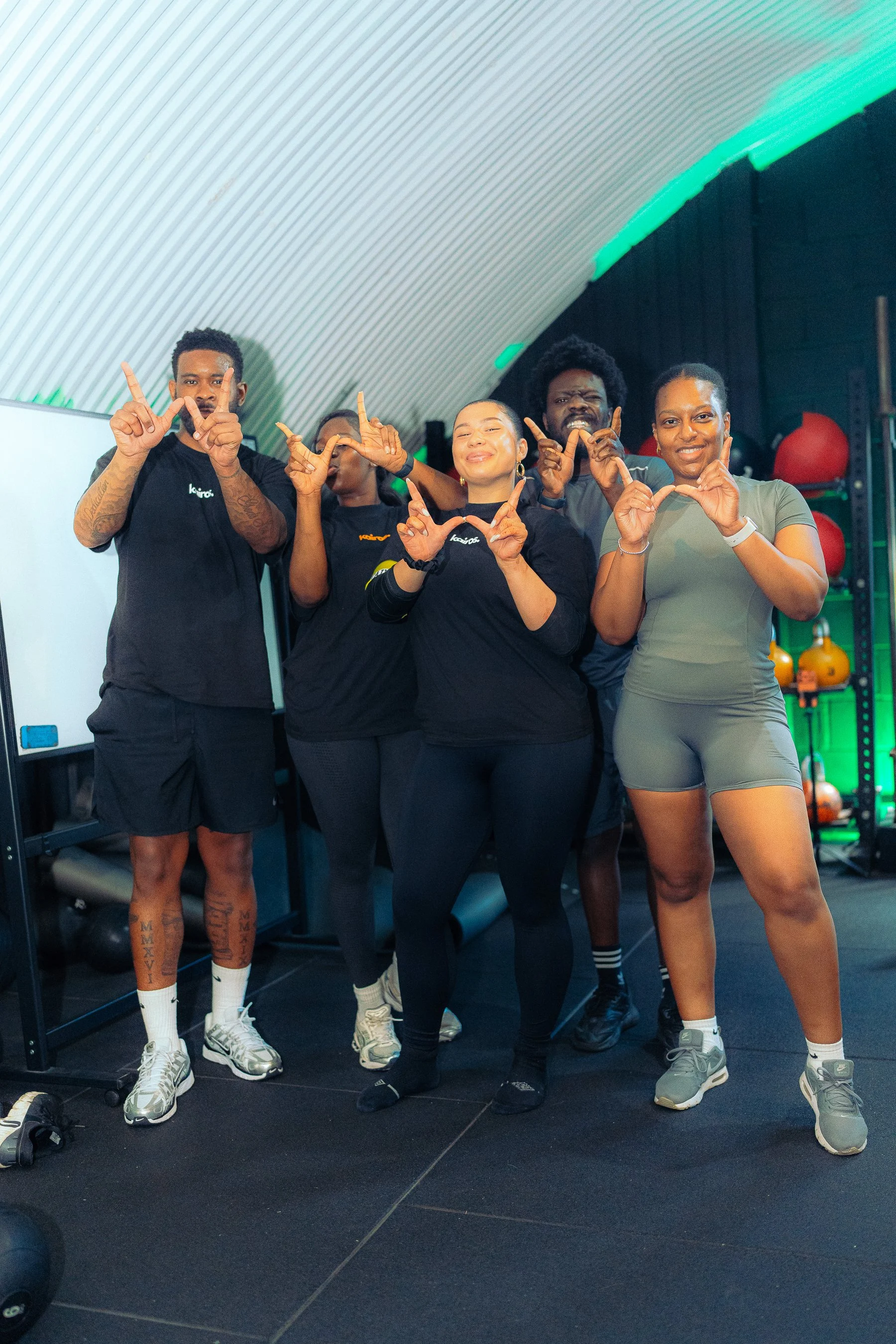 Group of five diverse people posing and making gestures in a gym with workout equipment and black walls.