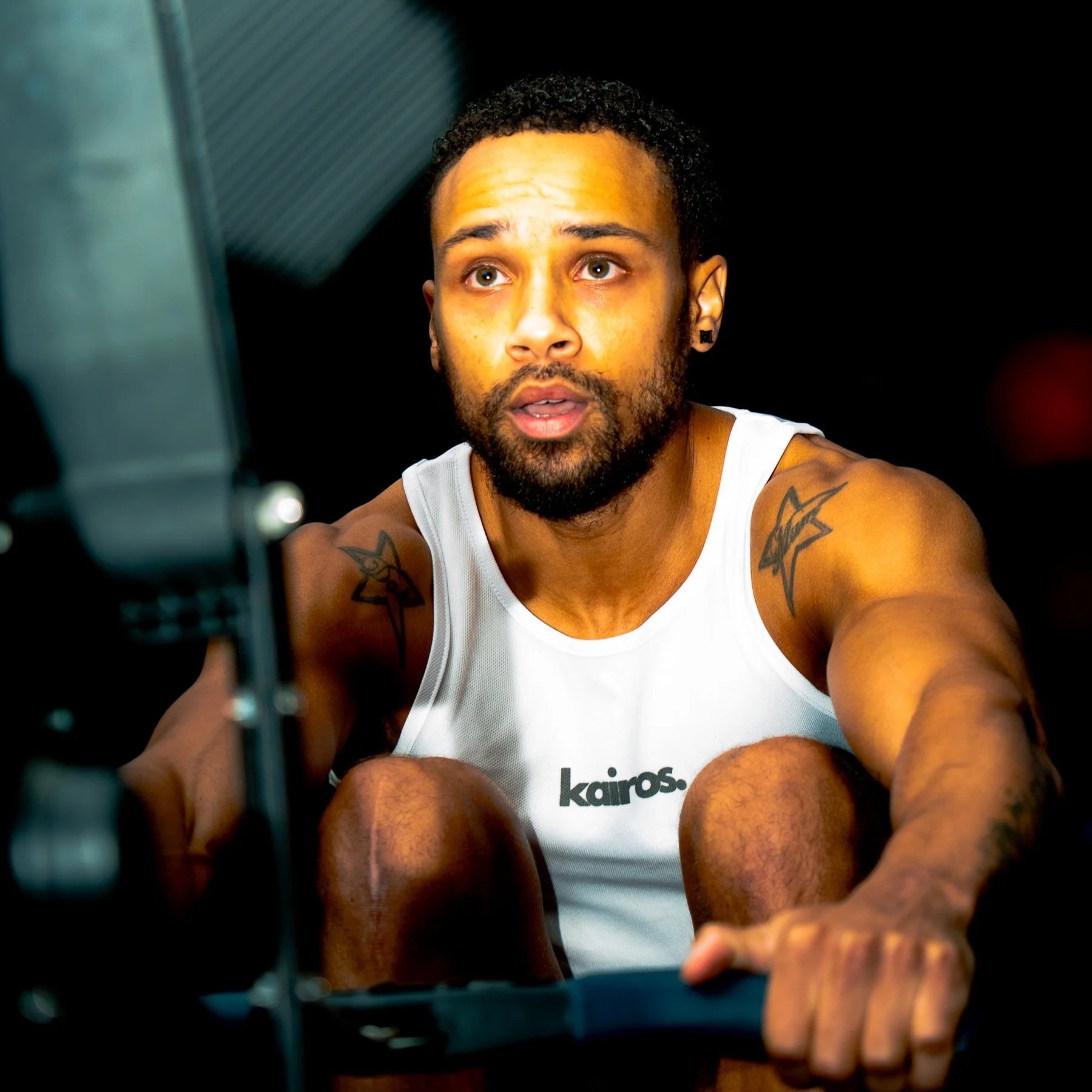 A man with short curly hair, beard, and tattoos on his arms, wearing a white tank top with the word Kairos on it, gazes at a camera with a serious expression, inside a dark room.