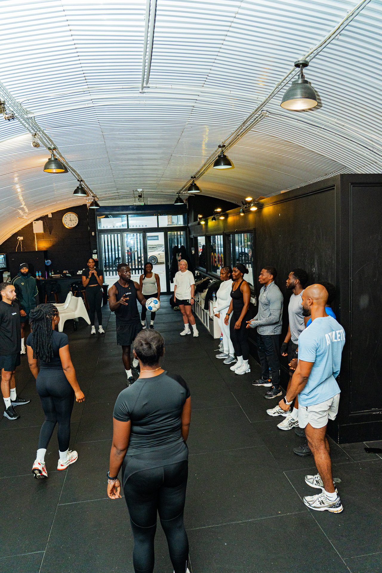 Group of people gathered in a gym, listening to an instructor demonstrating with a soccer ball.