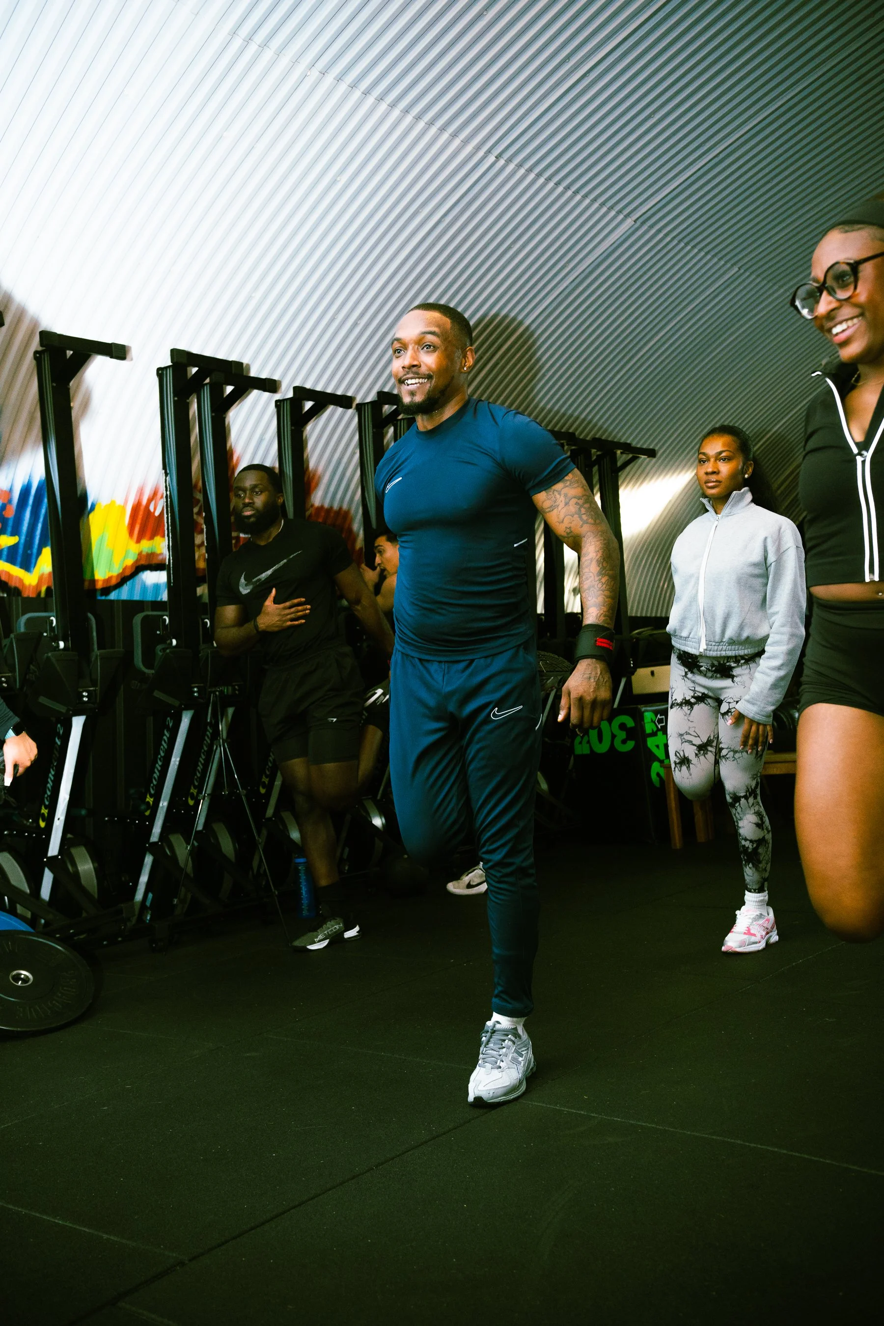 Group of four people in a gym, one man in a blue shirt and black pants exercising, three women watching, one woman in black with glasses, and another in gray with patterned leggings, all standing on a black gym floor.