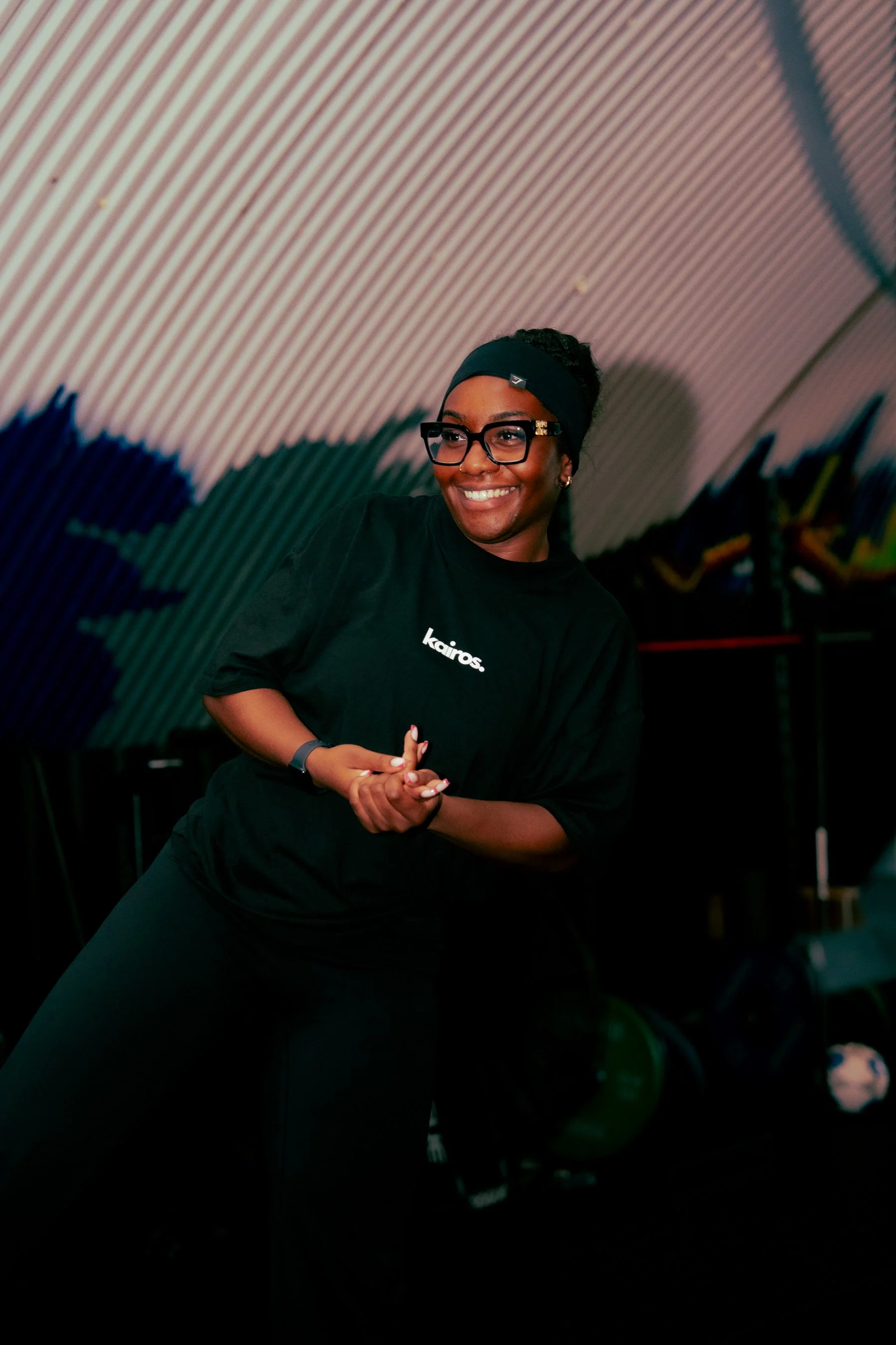 A woman smiling, wearing glasses, a dark headband, and a black shirt with the word 'kairos' written on it, standing in a room with a curved, ribbed ceiling and some plants in the background.