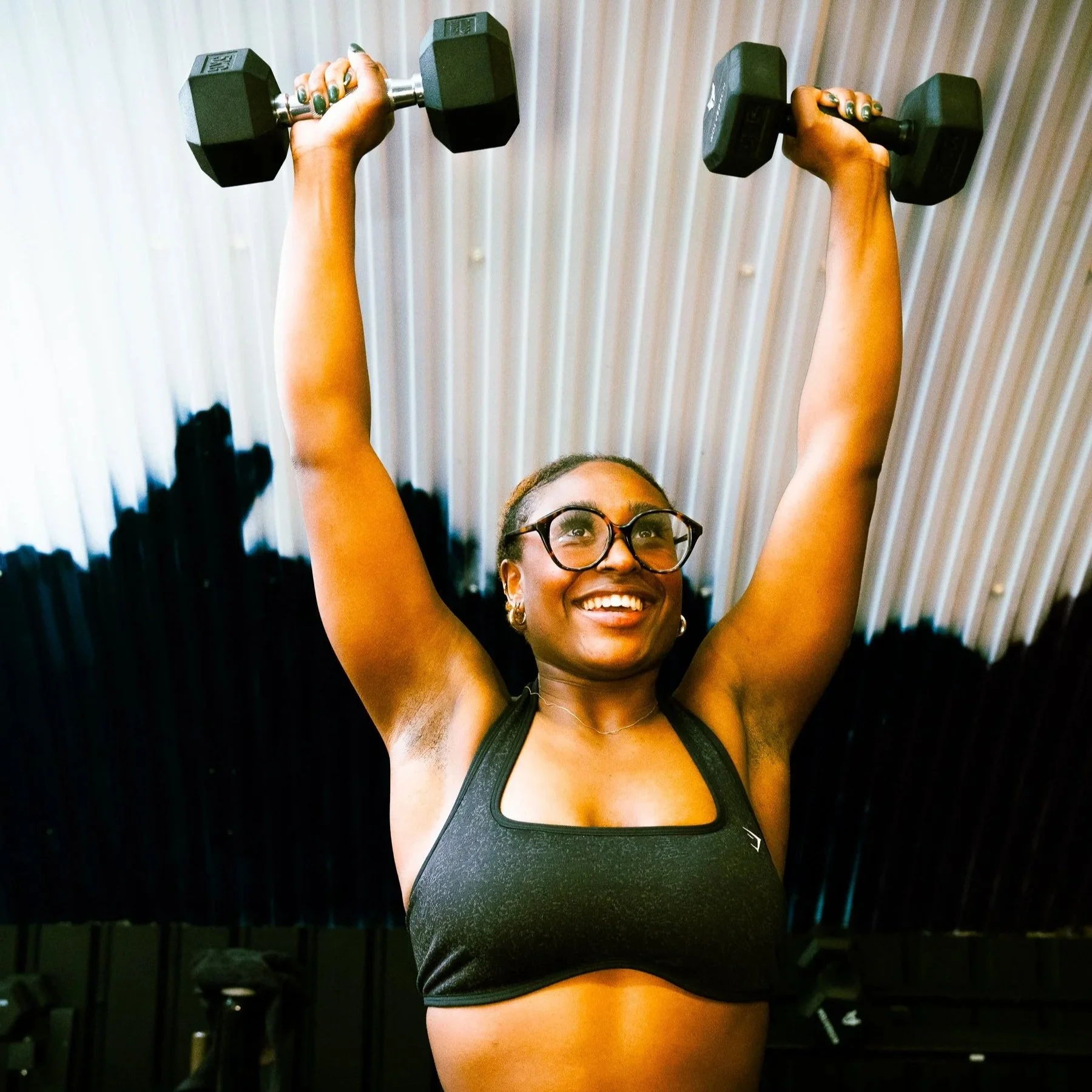 A woman with glasses smiling and lifting two dumbbells above her head in a gym or workout space with a corrugated ceiling.