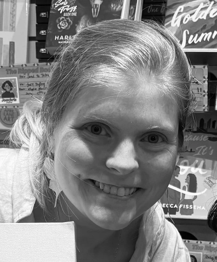 A woman with light-colored hair smiling indoors, with books and signs in the background.