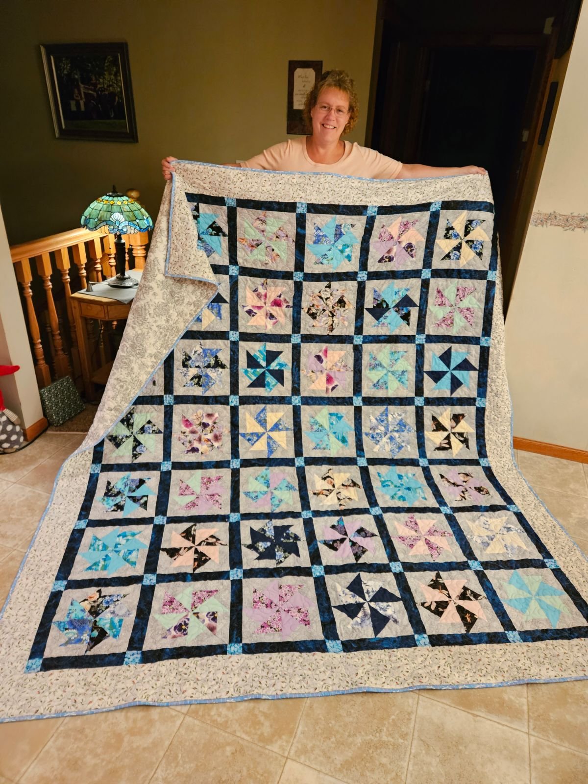 A woman smiling and holding a large colorful quilt with pinwheel and floral patterns, standing in a cozy home interior. The quilt features various pastel colors and blue accents, with a decorative border.