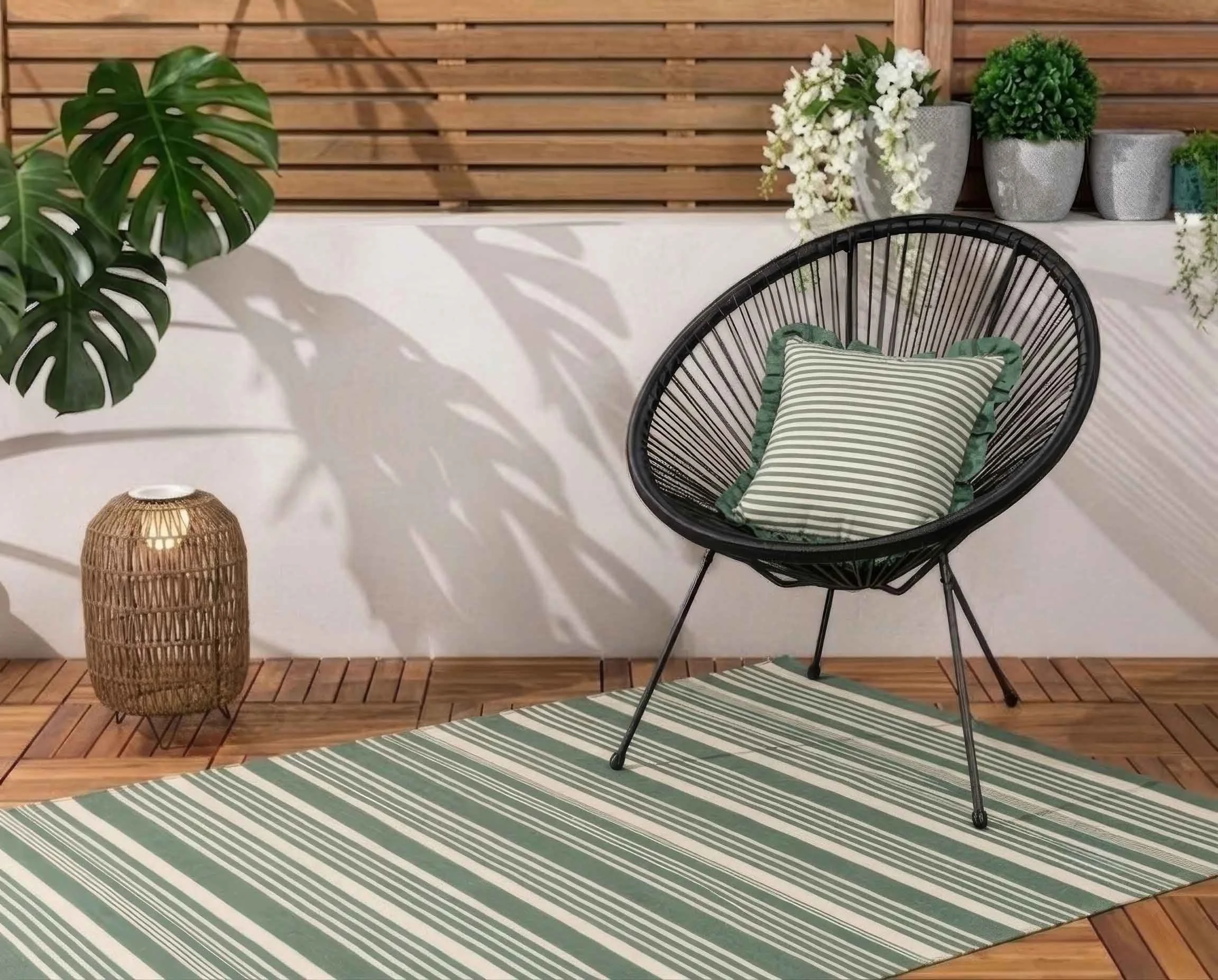 Outdoor patio area with a round black wire chair with a striped cushion, a woven lamp, and light green striped rug. A white wall with a wooden slat fence decorated with potted plants and white flowers.