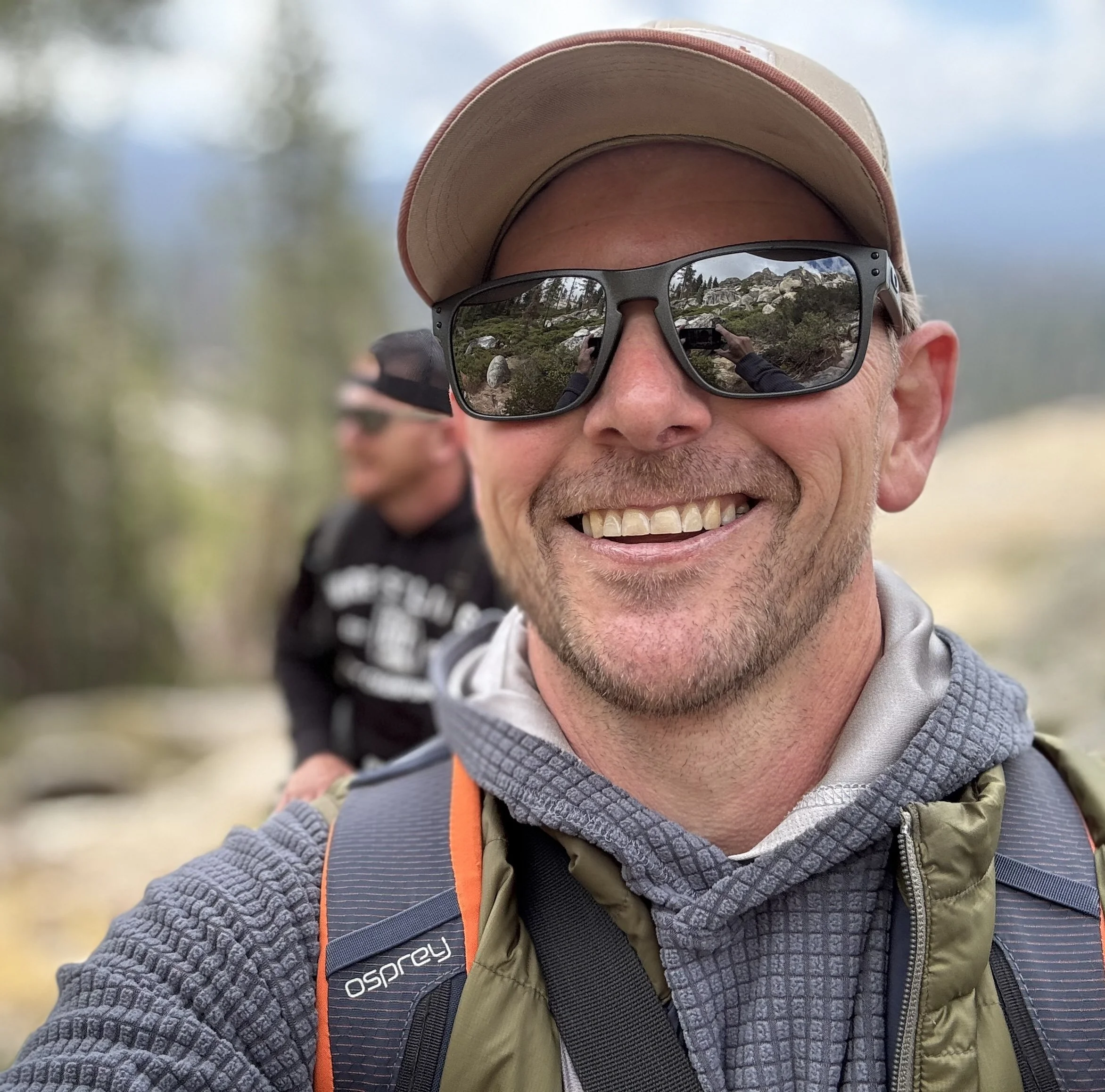 Close-up of a man wearing sunglasses, a beige cap, and outdoor clothing. Reflection in sunglasses shows rocky terrain and greenery.