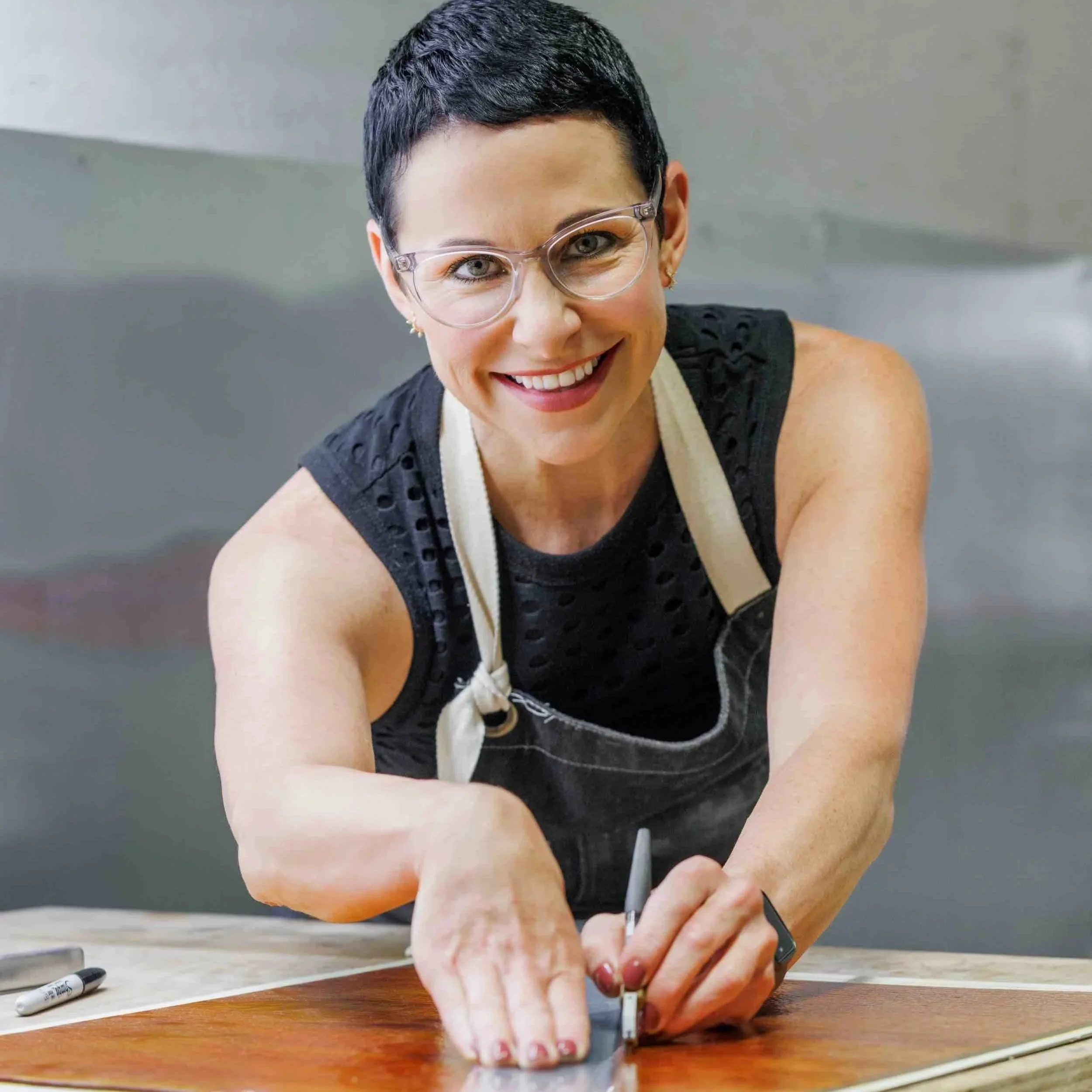 Woman with short dark hair, glasses, and a bright smile working on a project at a wooden table, with markers nearby.