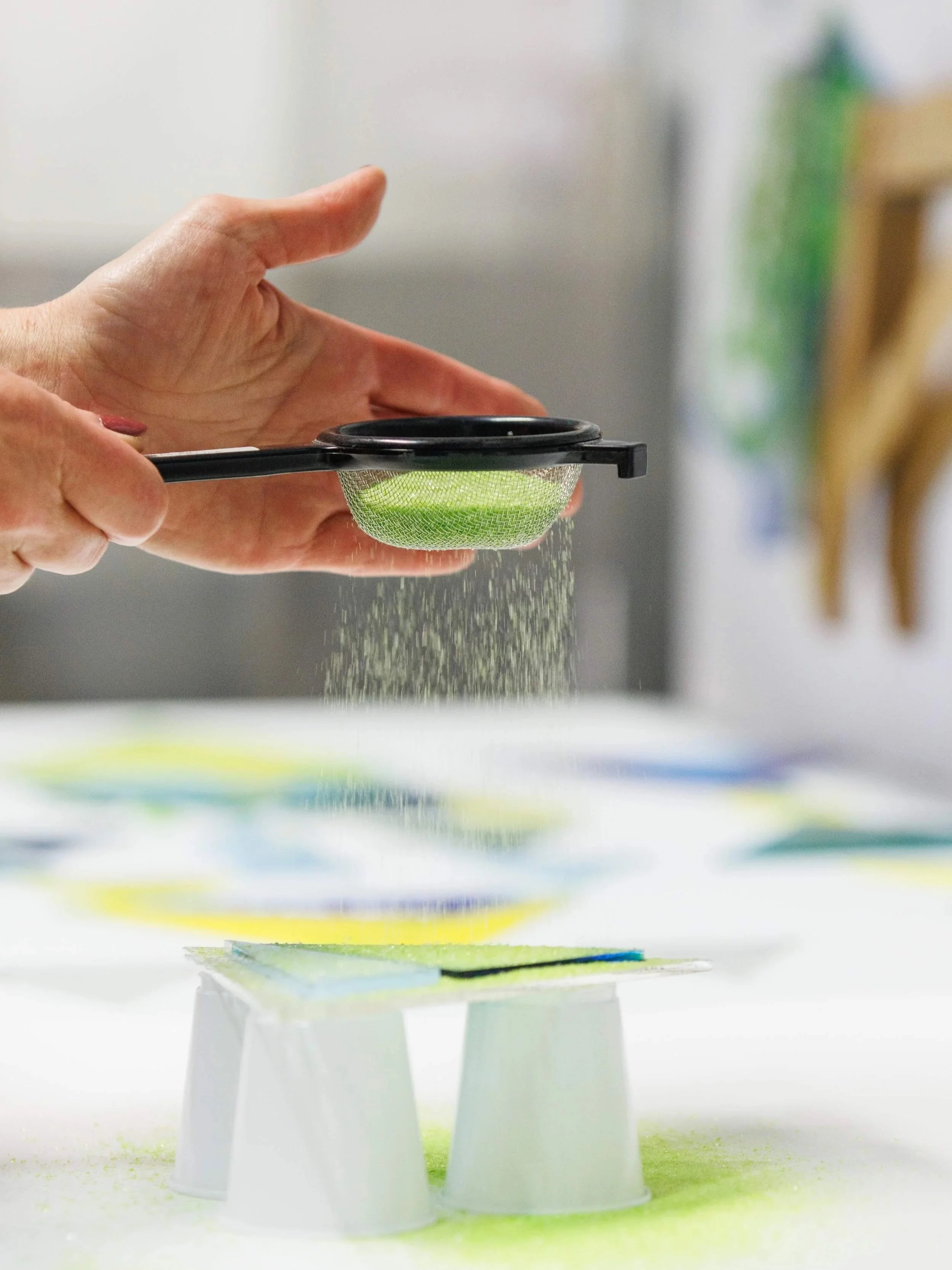 Fused glass artist sifting Spring Green Bullseye glass powder onto a glass sheet, creating a glass panel in her Atlanta studio.