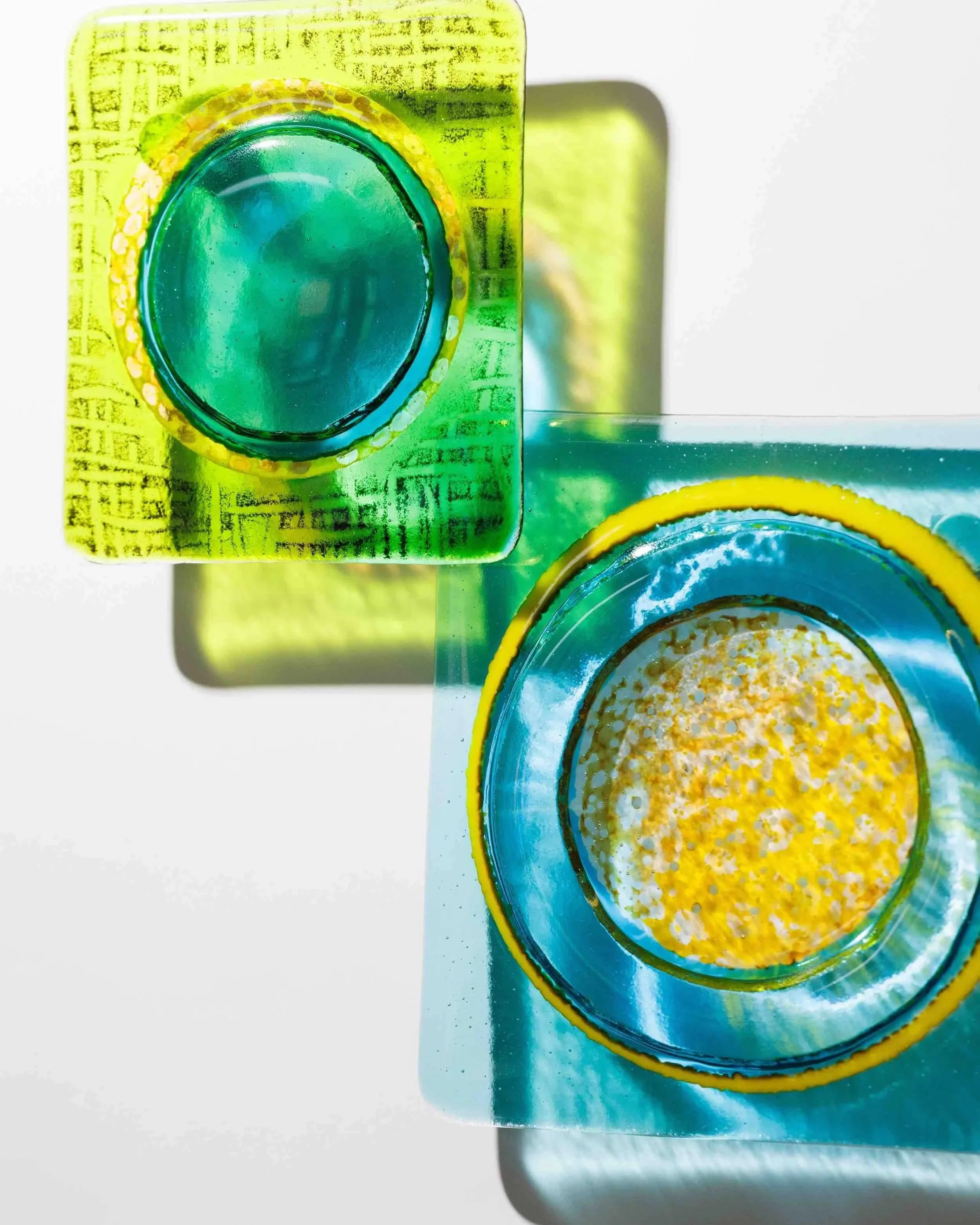 Close-up of colorful glass squares, mainly green and blue, with one yellow edge, placed on a white surface. The squares have a translucent appearance and textured details.