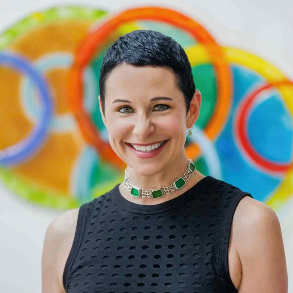 A woman with short black hair, smiling, wearing a black sleeveless top and a green gemstone necklace, with colorful abstract art in the background.