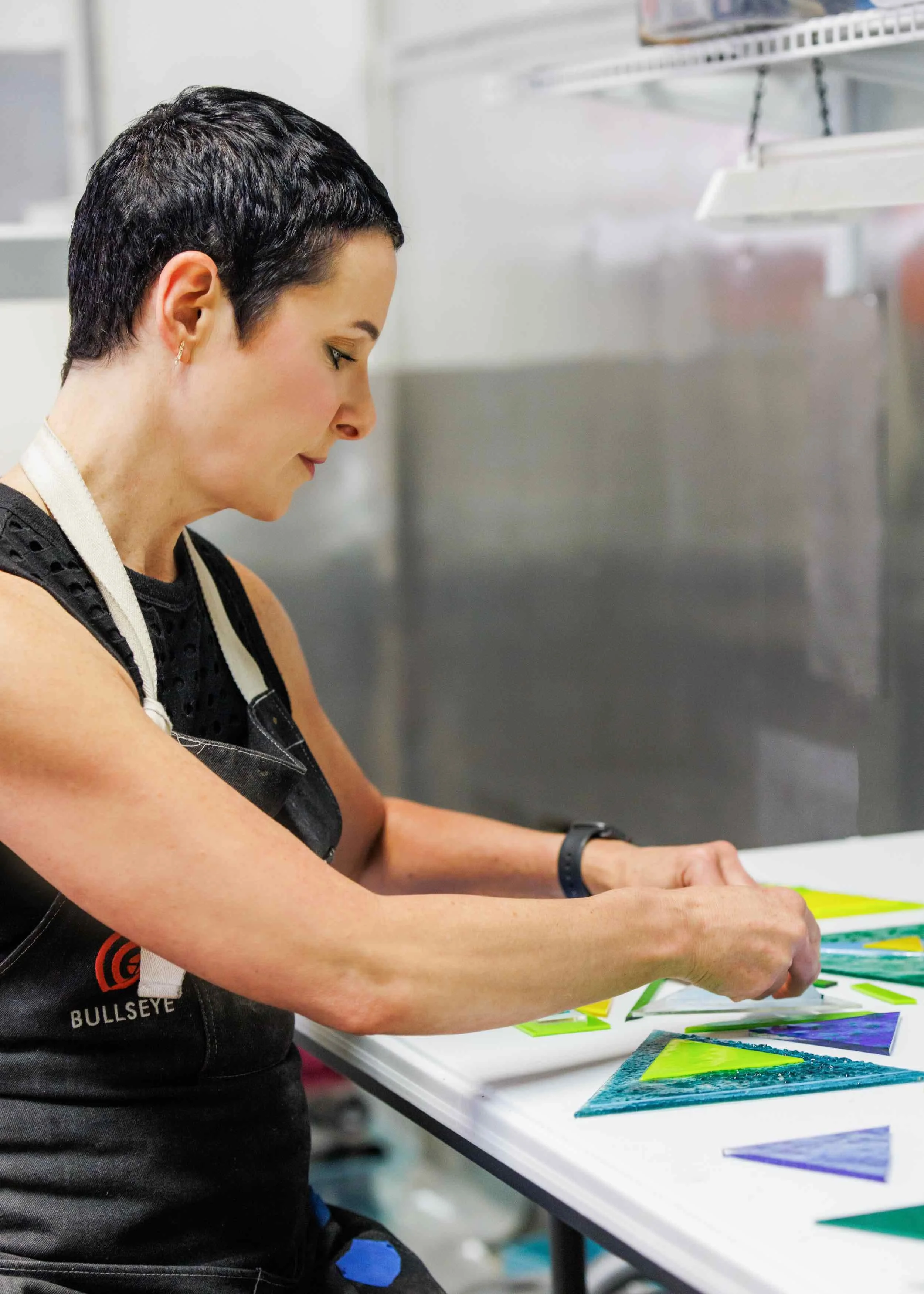 A woman with short dark hair and earrings is working on a colorful art project on a table. The project involves bright, geometric shapes with textured and patterned details, and she appears focused on her work.