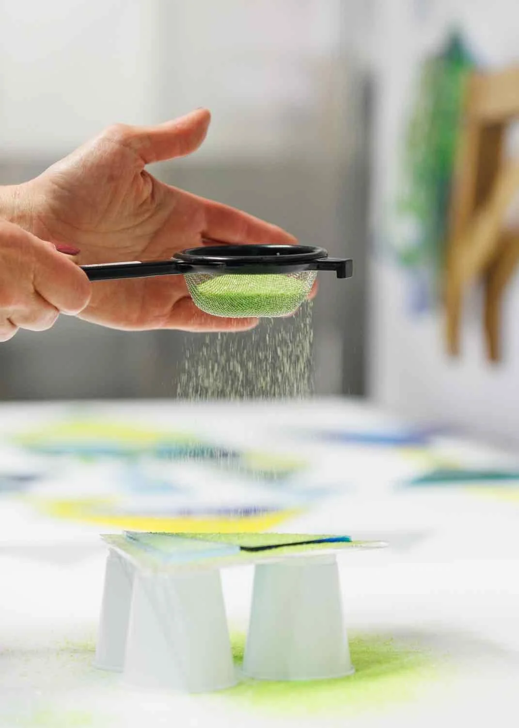 A person sifting colorful powder through a green mesh strainer over a table with paper and small white cups in a bright room.