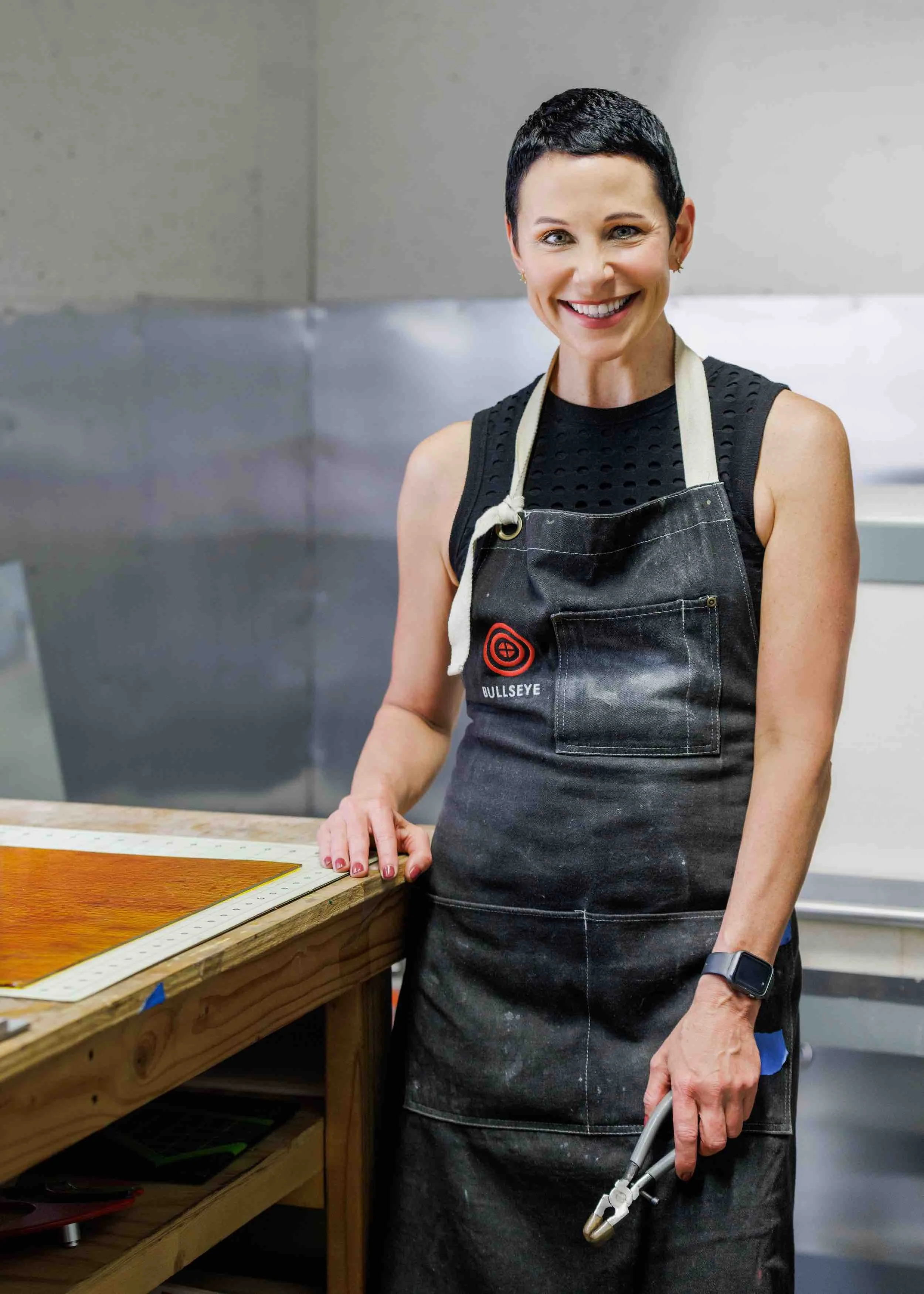 A woman with short dark hair smiling wearing a black sleeveless top and apron, standing in a workshop or kitchen, holding a tool in her right hand.